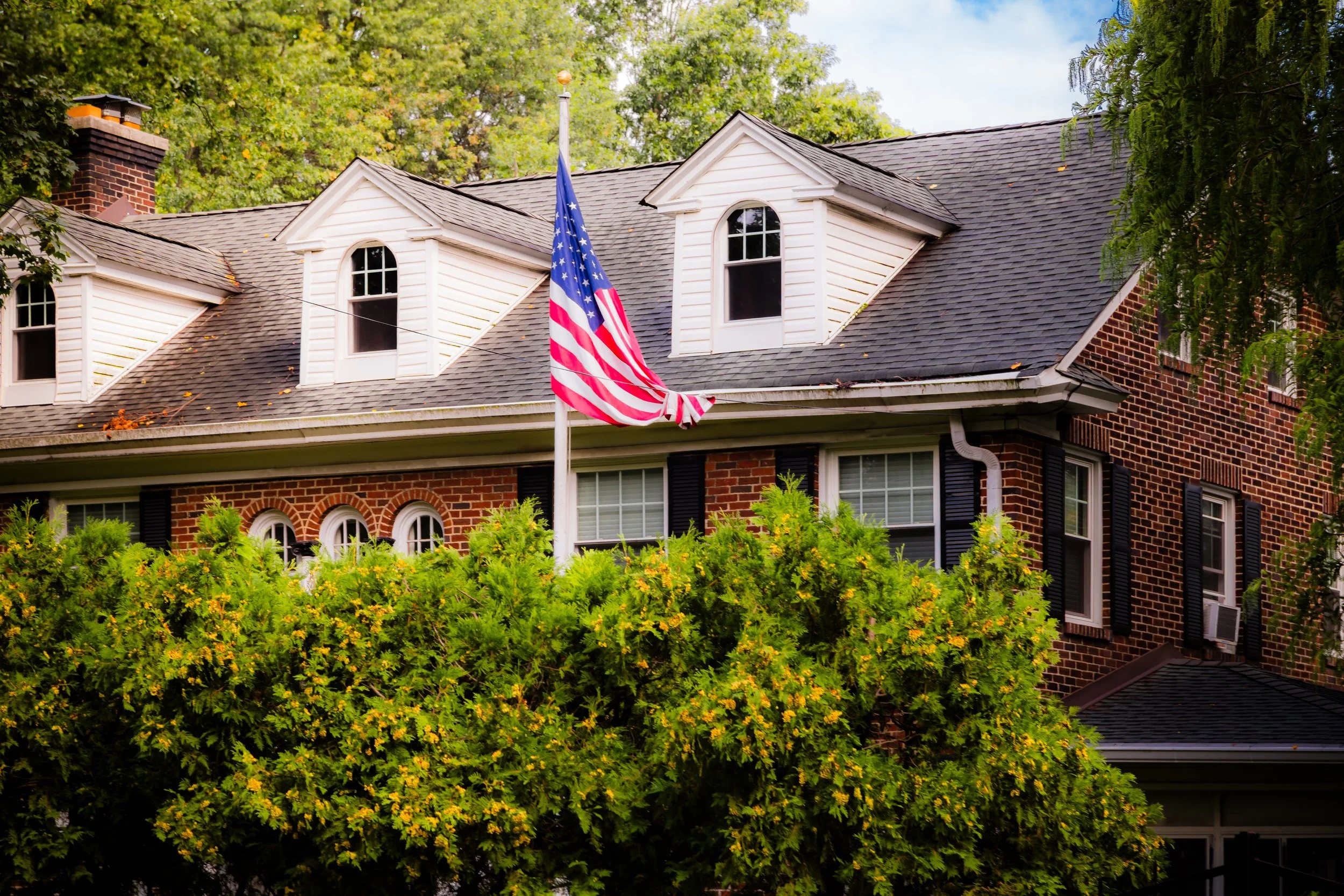 A brick house with white dormer windows and a gray shingle roof. An American flag is mounted on a pole in front of the house, surrounded by green trees and bushes.