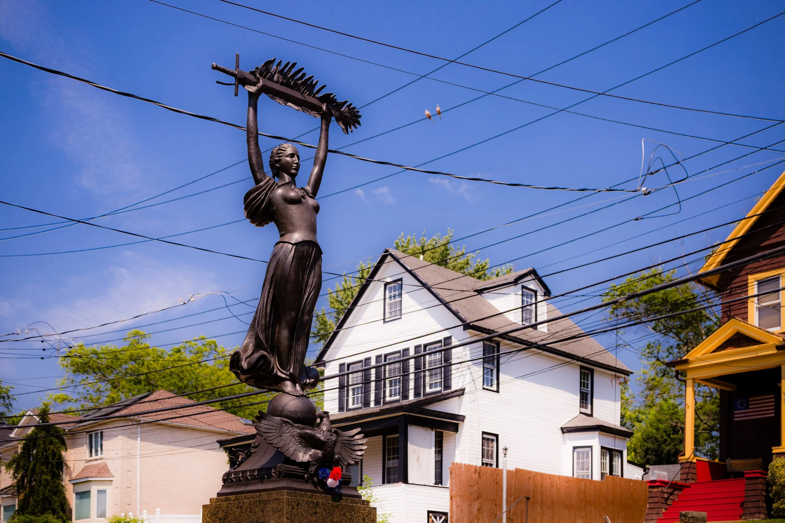 A bronze statue of a woman holding a rake, standing on a pedestal with an eagle at her feet, in front of houses and power lines under a blue sky.
