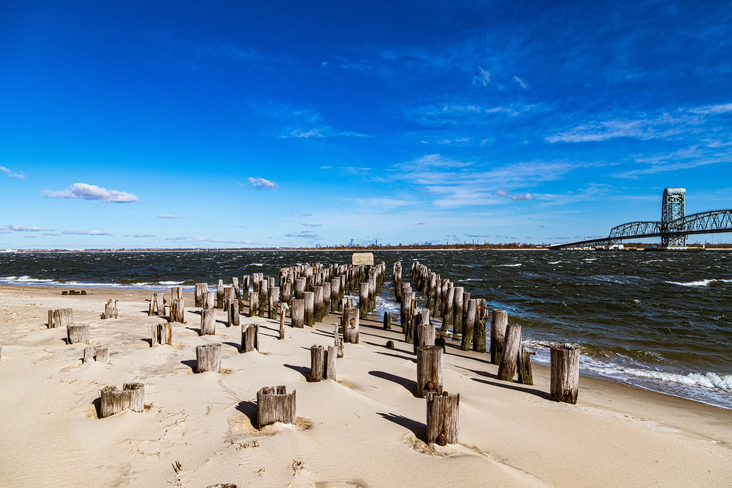 A sandy beach with wooden posts leading into the water, a bridge in the background, and a blue sky with scattered clouds.