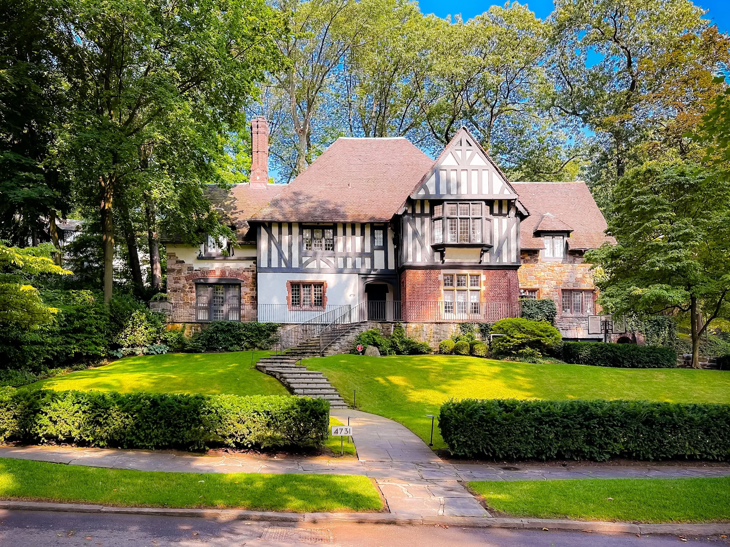 A large, historic house with Tudor-style architecture, featuring a mix of brick, stone, and half-timbering, surrounded by a lush green lawn and trees.