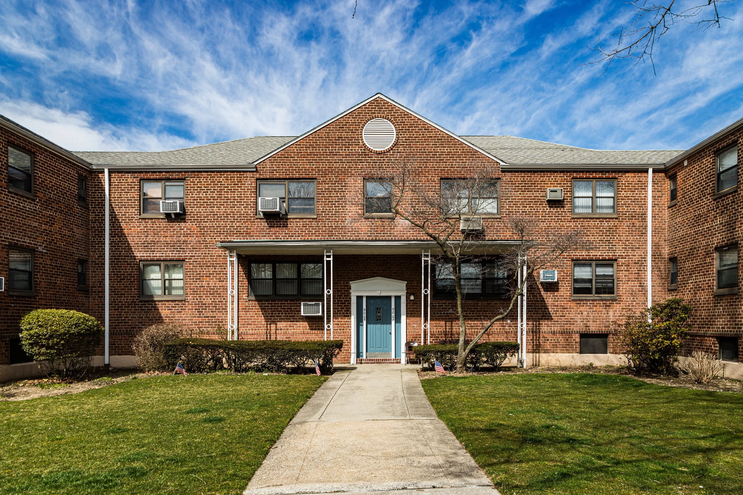 A brick apartment building with a front entrance, a paved walkway, and fenced shrubs. The building has multiple windows with window air conditioning units, a central round vent, and a blue door. The lawn is well-maintained with trees and small flags.