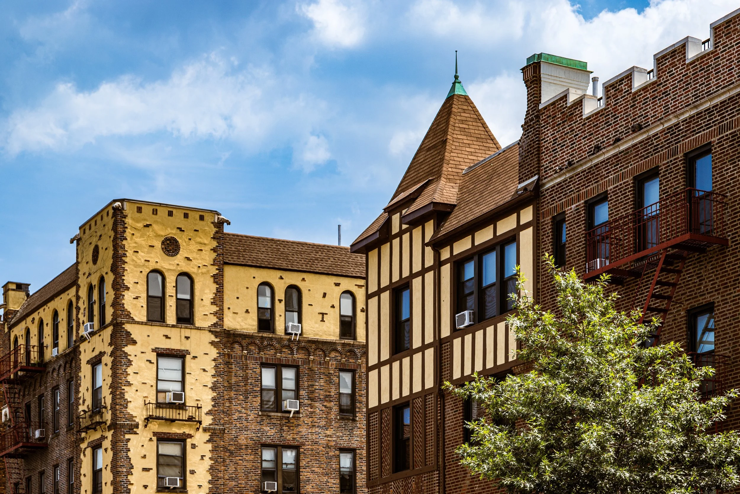 Older urban buildings made of brick and stucco with decorative features, fire escape, and a tree in front, under a partly cloudy sky.
