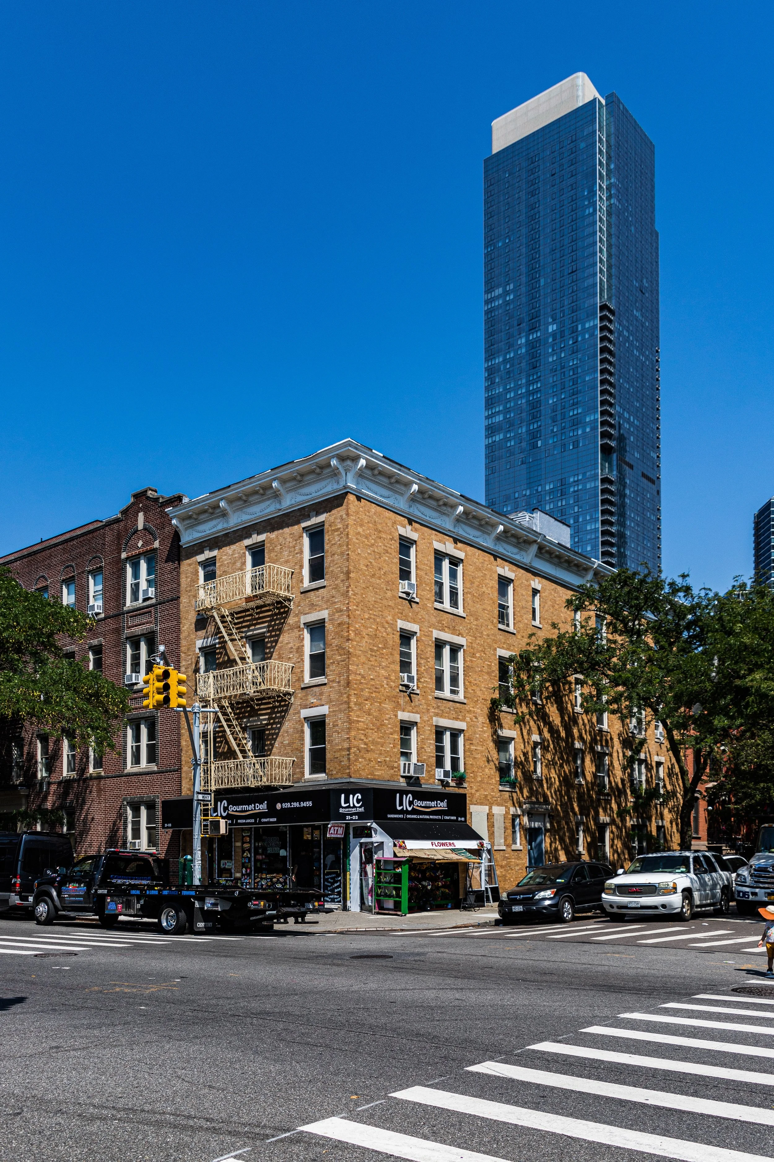 A city street scene with a three-story brick building with a grocery store on the ground floor and a tall modern skyscraper in the background. There are cars parked along the street, a crossing, trees, and a traffic light.