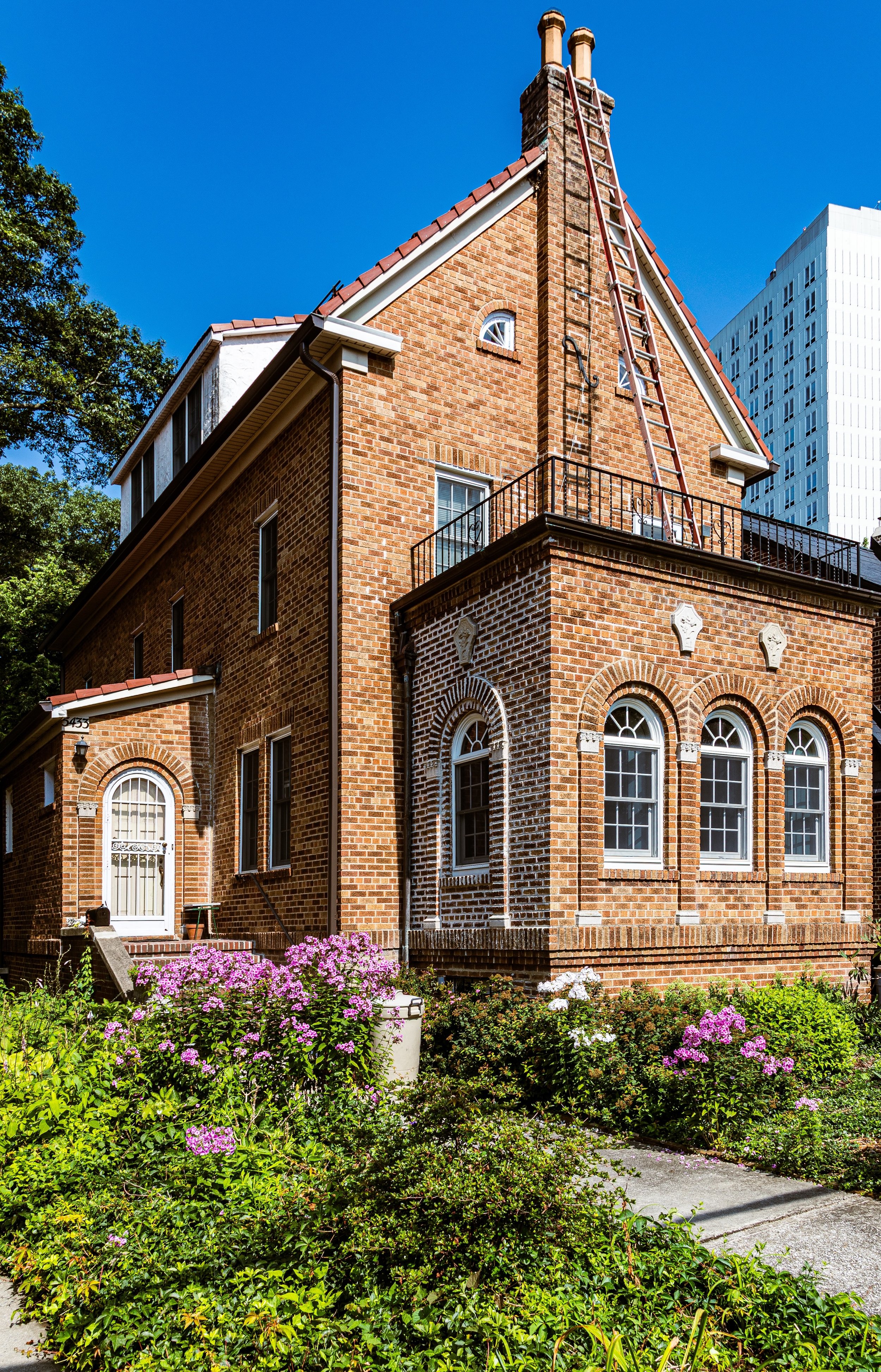 A brick multi-story house with arched windows and a garden with pink and white flowers in the foreground, with a ladder resting against the chimney on a clear blue sky day.