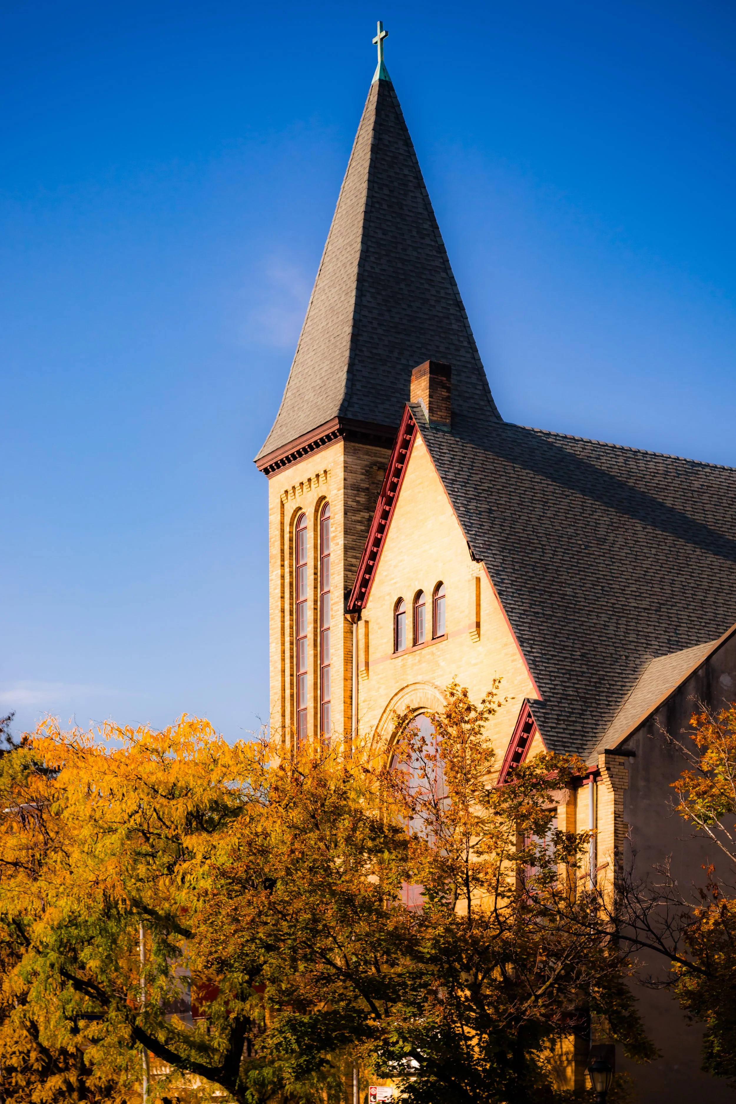 A church with a tall steeple topped with a cross, surrounded by trees with fall foliage, under a clear blue sky.