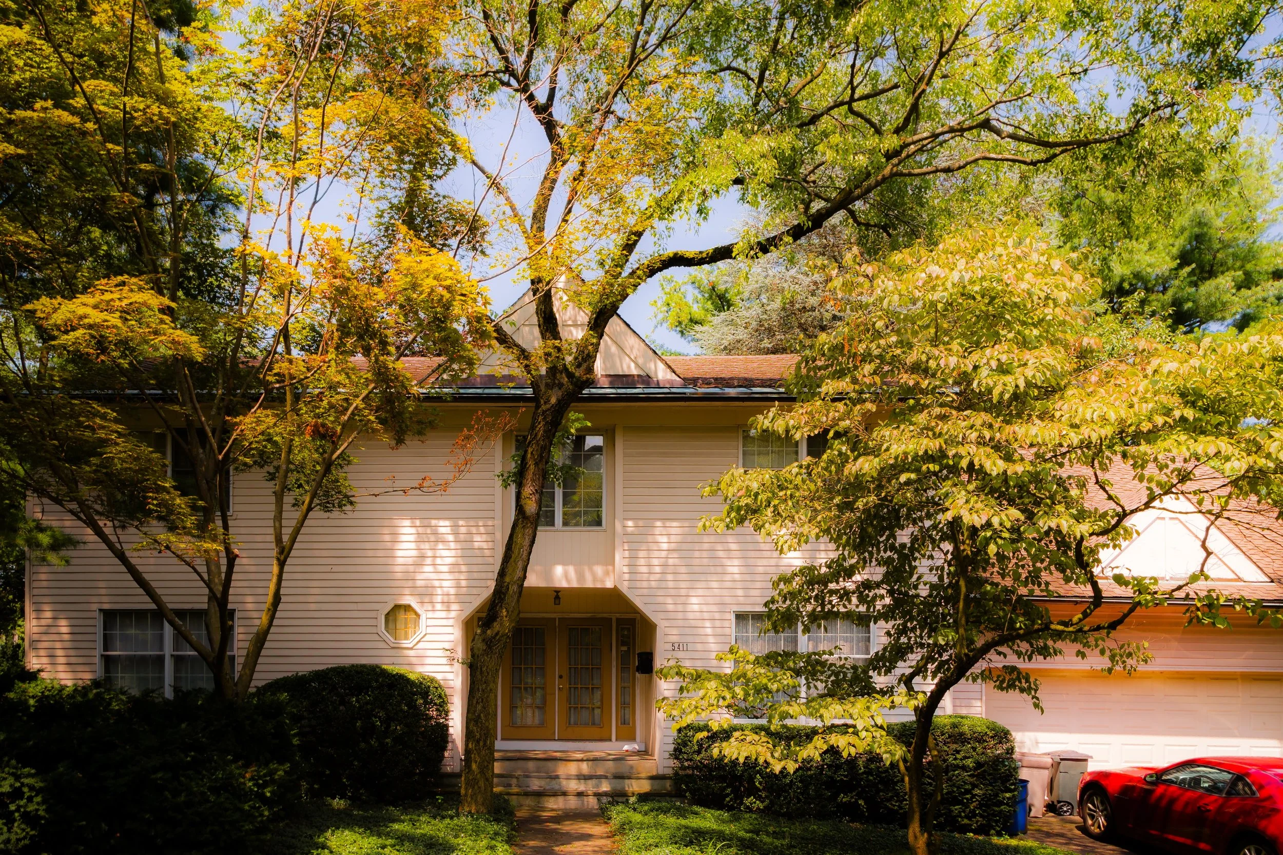 A two-story house with beige siding, a brown roof, and a porch. The house is surrounded by trees with green and yellow leaves and a green lawn. A red car is parked in the driveway on the right.