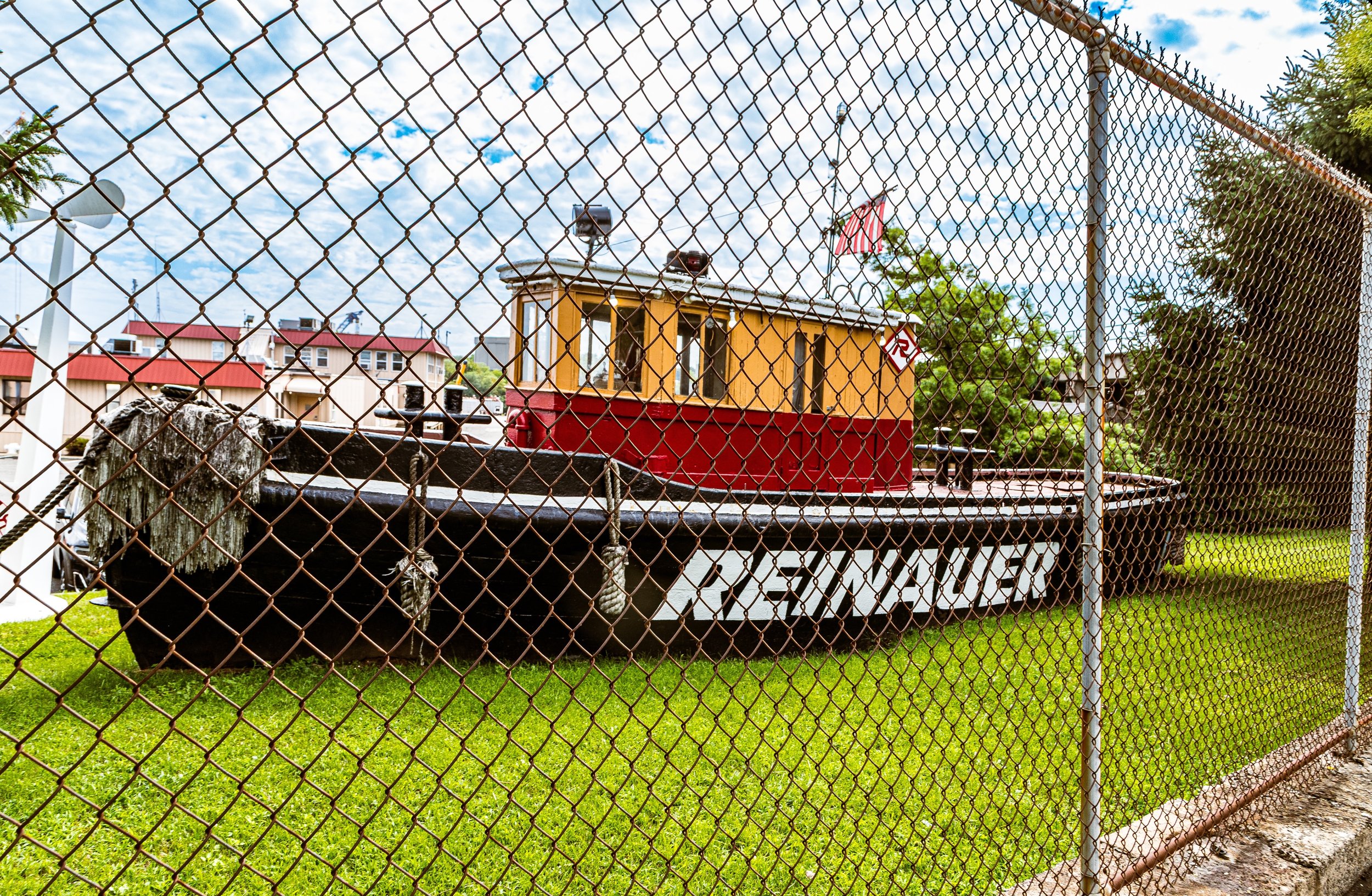 A boat called the USS Voyager behind a chain-link fence with a grass lawn in the foreground and buildings in the background.