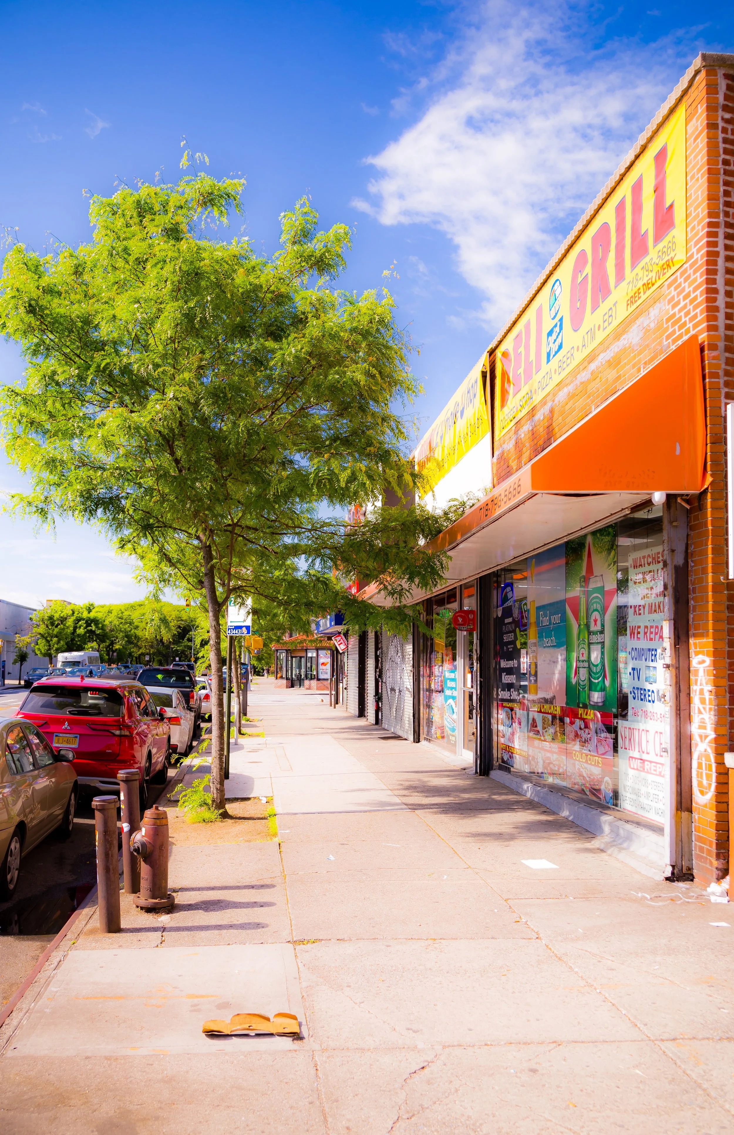 A sidewalk with parked cars, green trees, and storefronts on a sunny day with a blue sky and white clouds.