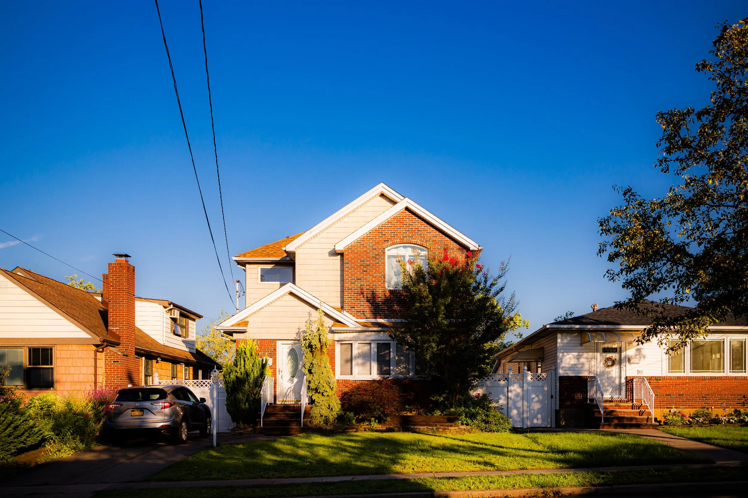 A sunny suburban neighborhood with three houses and a blue sky. The houses have brick and siding exteriors, steps leading to their front doors, and well-maintained lawns with shrubs and trees.