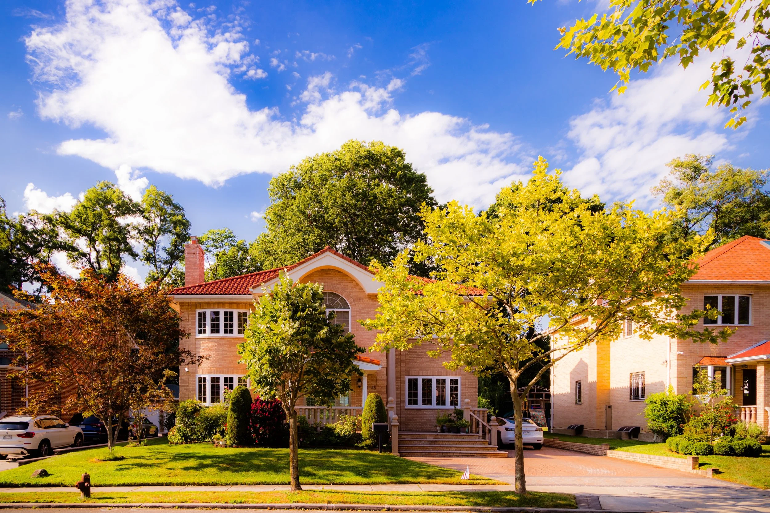 A sunny suburban street with large trees, a two-story brick house with a red-tile roof, and a well-maintained lawn.