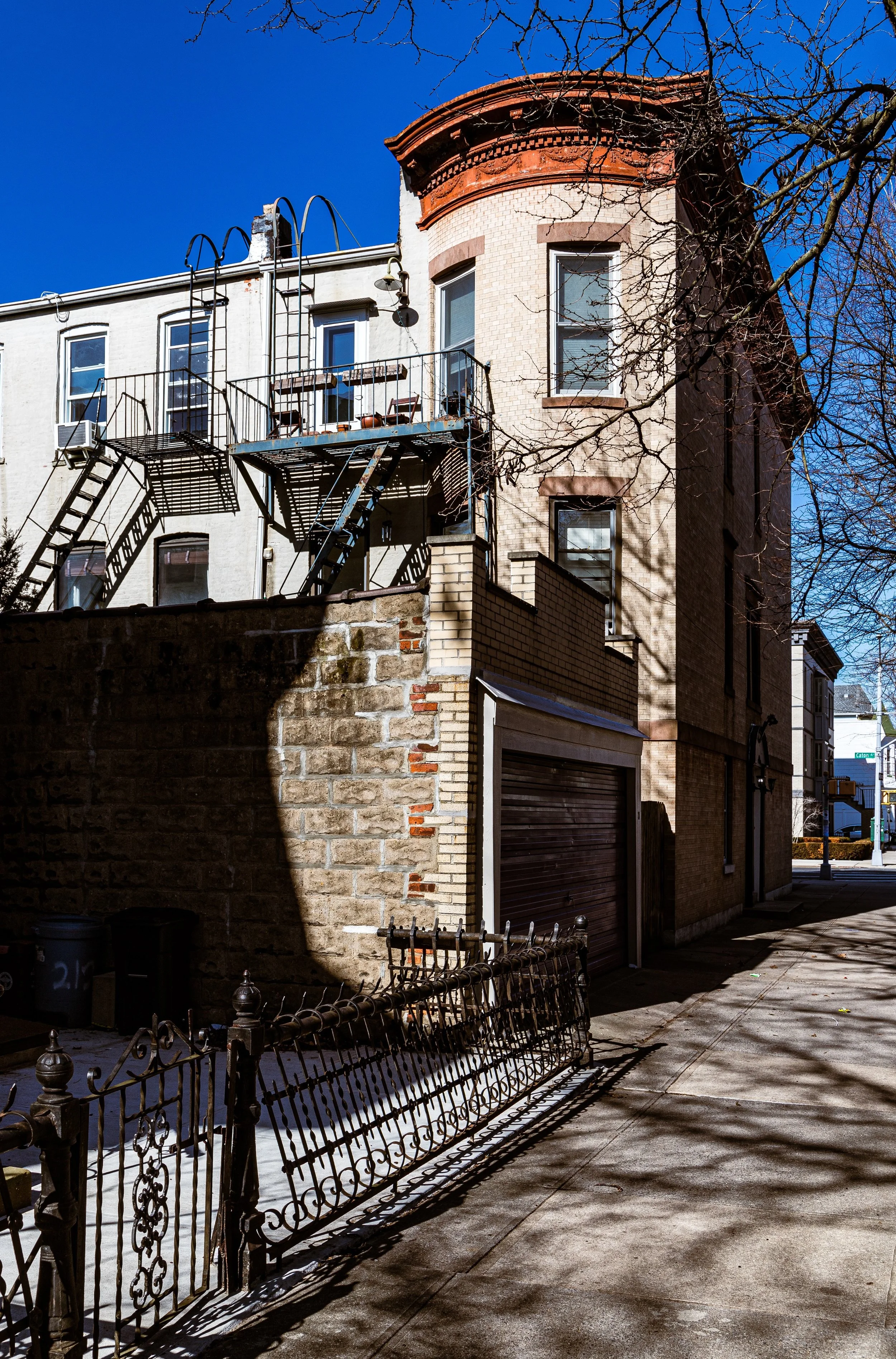 An urban building corner with a brick wall on the ground floor and a beige upper story. There is a fire escape with stairs and a small balcony. Shadows of tree branches are cast on the sidewalk and building, under a clear blue sky.