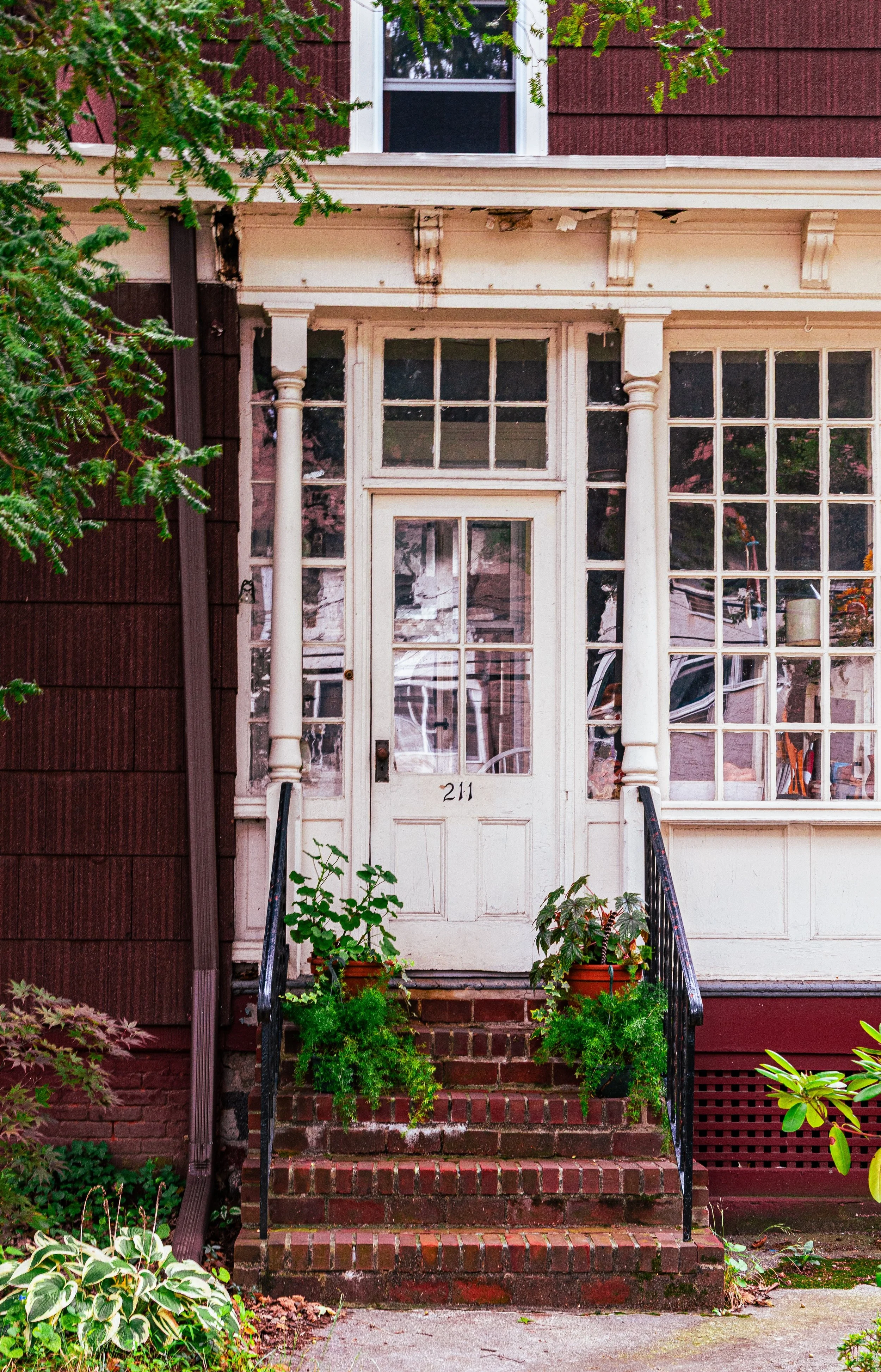 Front entrance of a house with brick stairs, white door with the number 211, and large windows. There are potted plants on the steps and greenery around.