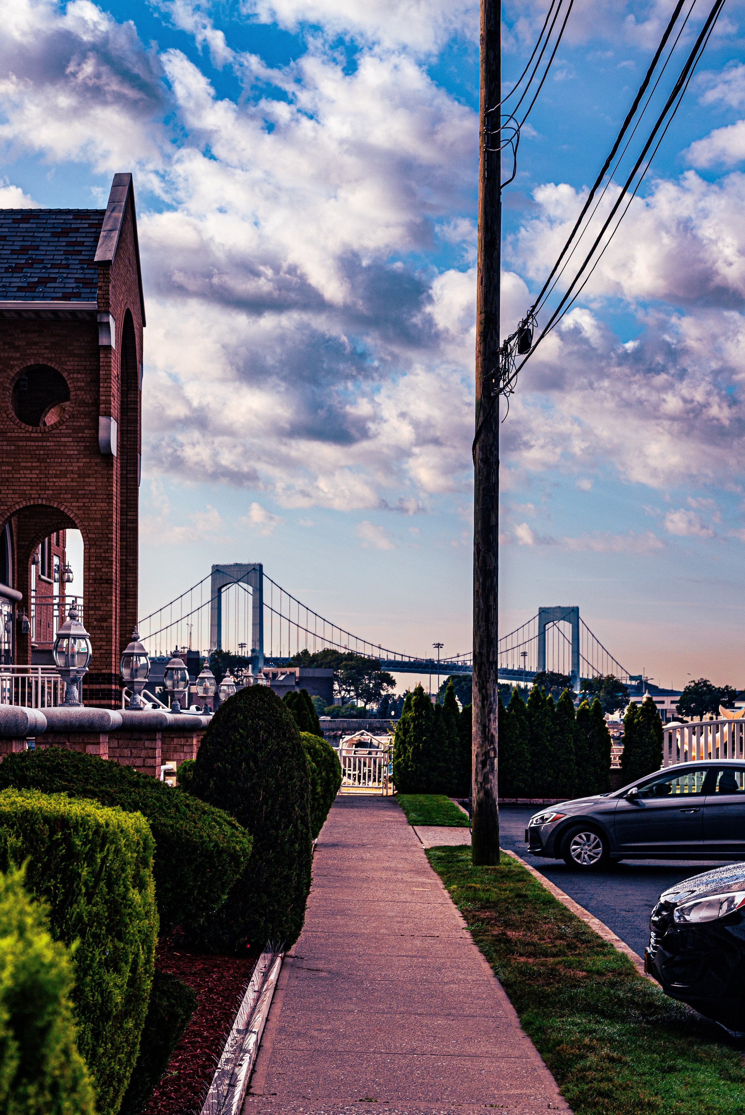 A sidewalk running through a fenced yard with shrubs and parked cars, with a bridge visible in the distance under a partly cloudy sky.