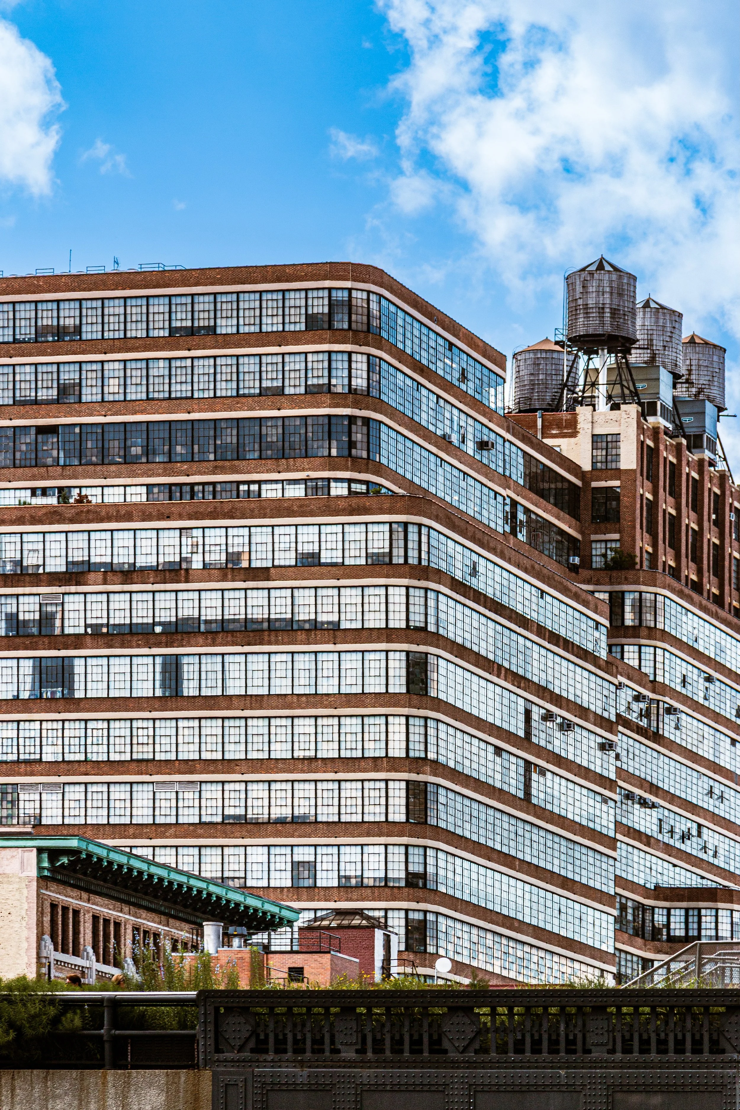 A large multi-story brick building with many windows, water towers on the roof, and a partly cloudy blue sky in the background.