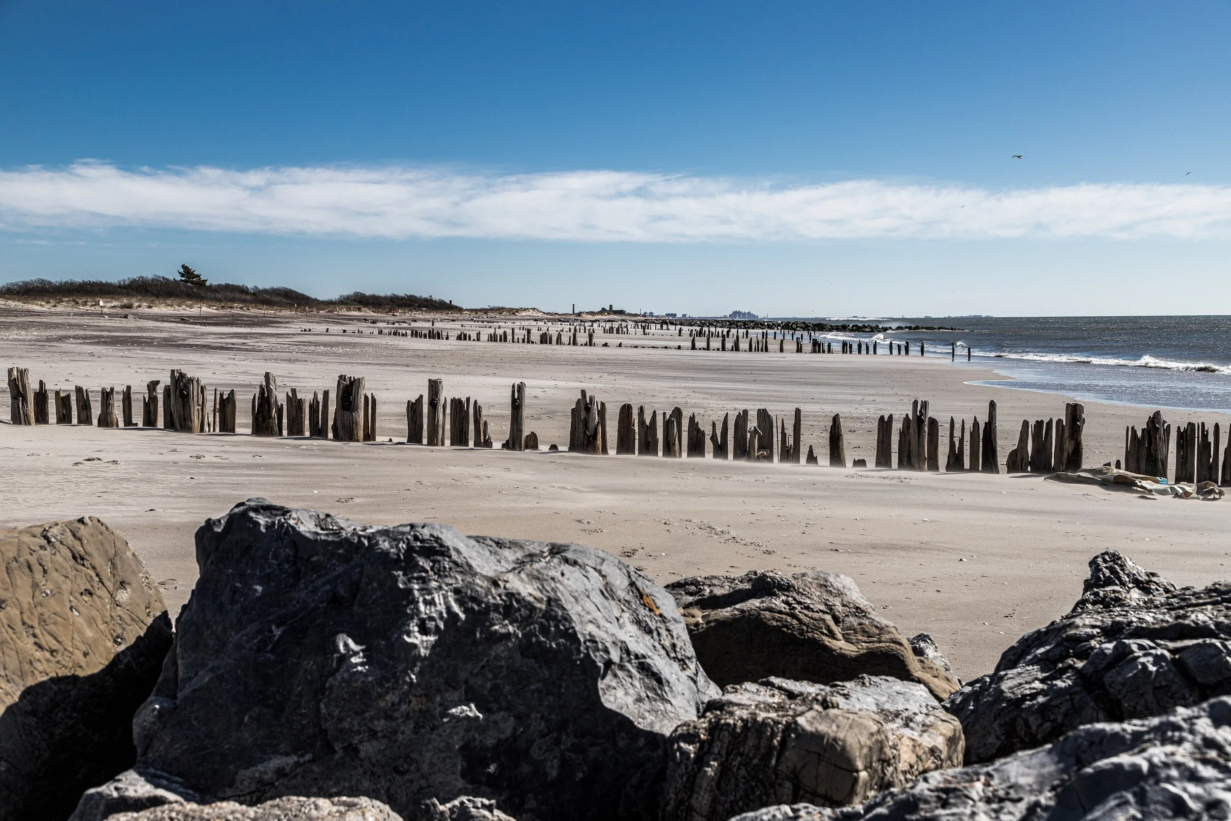 A sandy beach with rocks in the foreground, weathered wooden posts along the shore, and a clear blue sky with a few clouds.