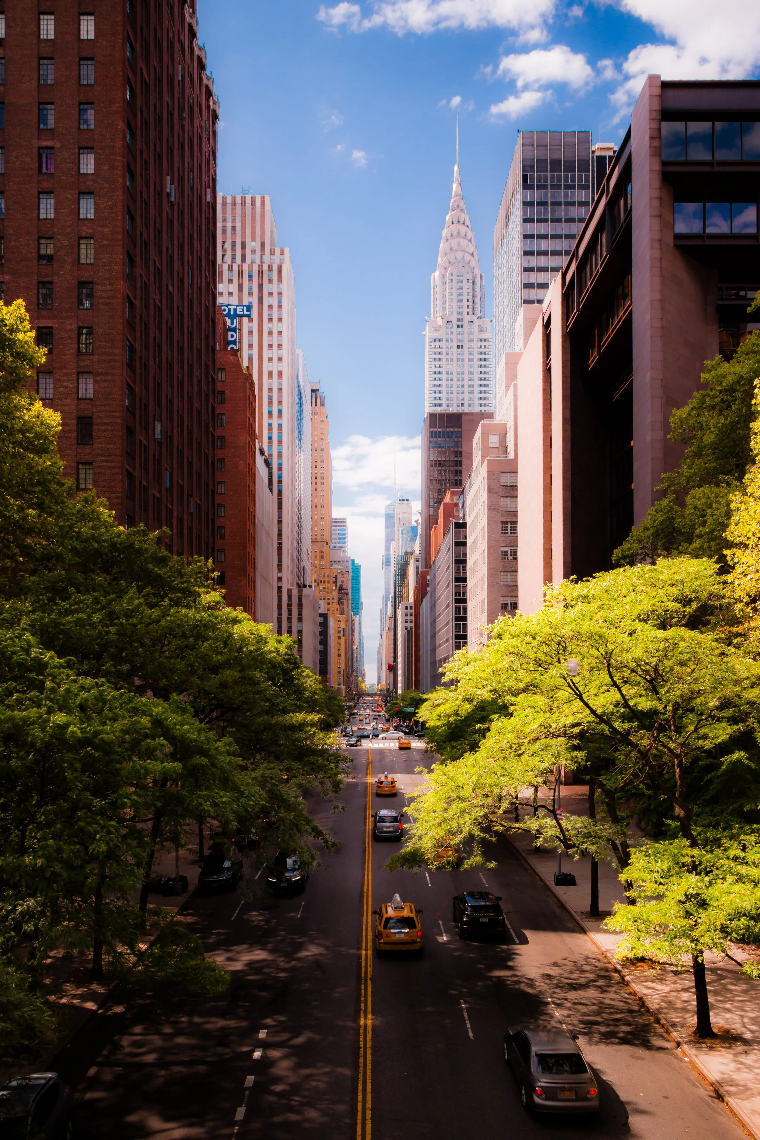 A city street scene with tall skyscrapers lining both sides, trees with green leaves in the foreground, cars and taxis on the road, and a partly cloudy blue sky overhead.