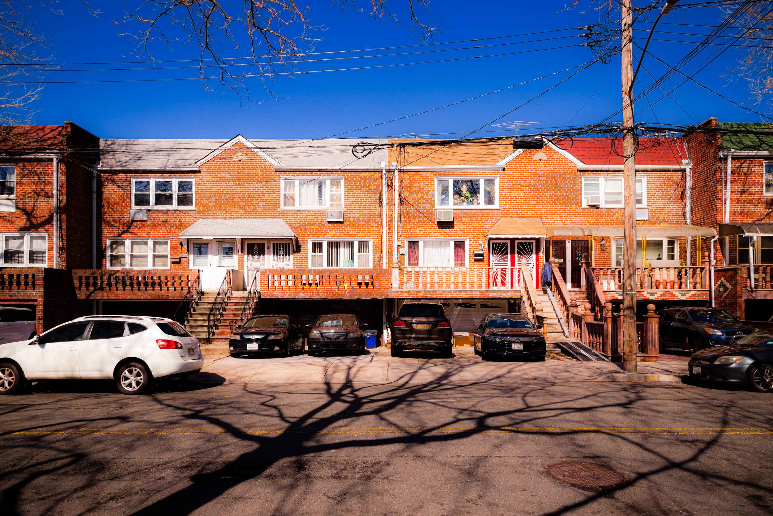 A row of five attached brick townhouses with stairs leading up to their entrances, cars parked in front, and a street with shadows of trees and a utility pole.