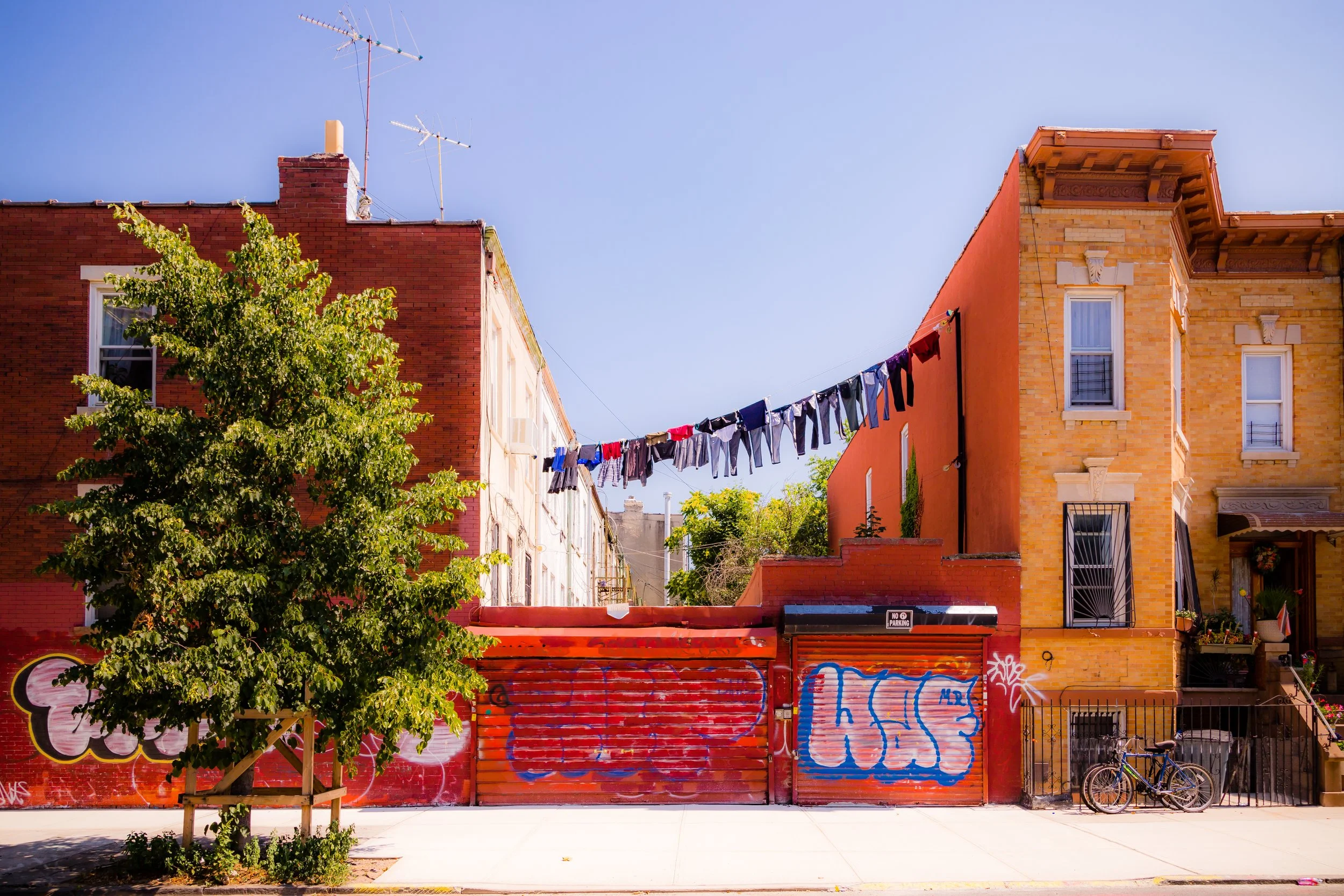 Colorful brick buildings with clothes hanging on a line, a tree in the foreground, bikes parked against the fence, graffiti on a red wall, and a clear blue sky.