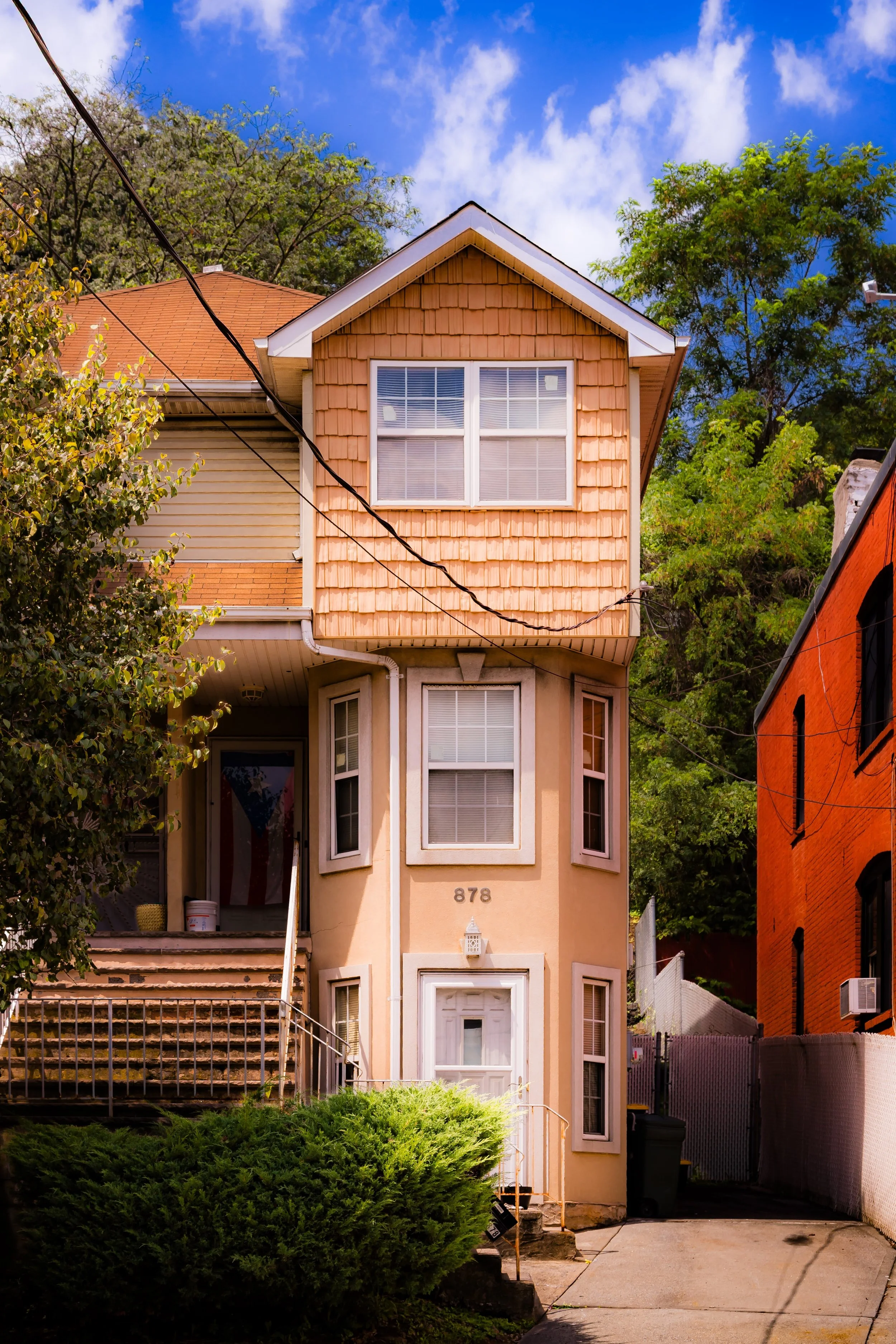 A narrow multi-story house with yellow and tan siding, multiple windows, and a bay window facing the front, with stairs leading up to the front door. The house has the number 878 near the door. There are trees, bushes, and neighboring houses visible around it, under a partly cloudy blue sky.