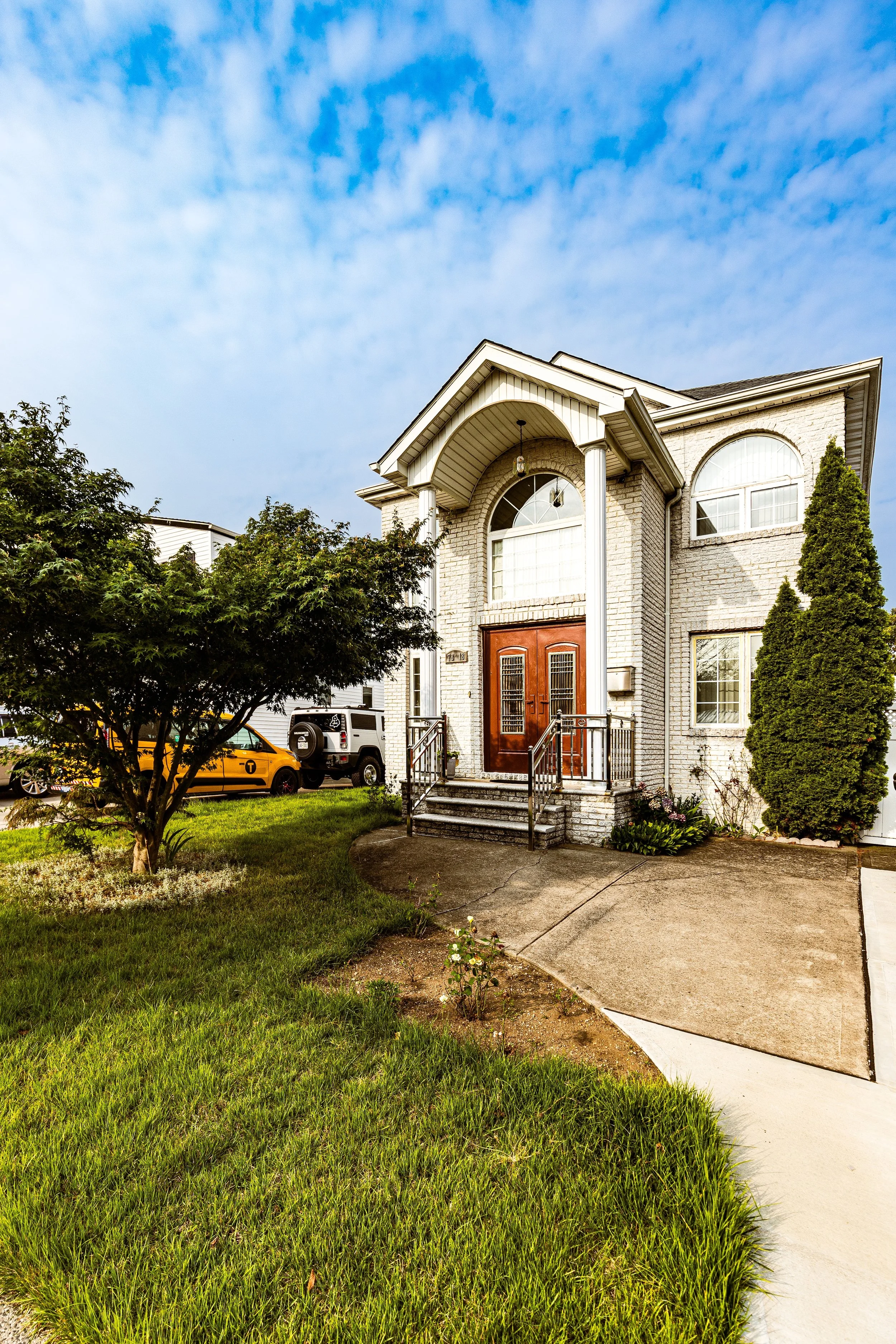 Front view of a two-story white brick house with a wooden front door, stairs with metal railings, a small porch, a large tree, and parked cars in the driveway, under a partly cloudy sky.