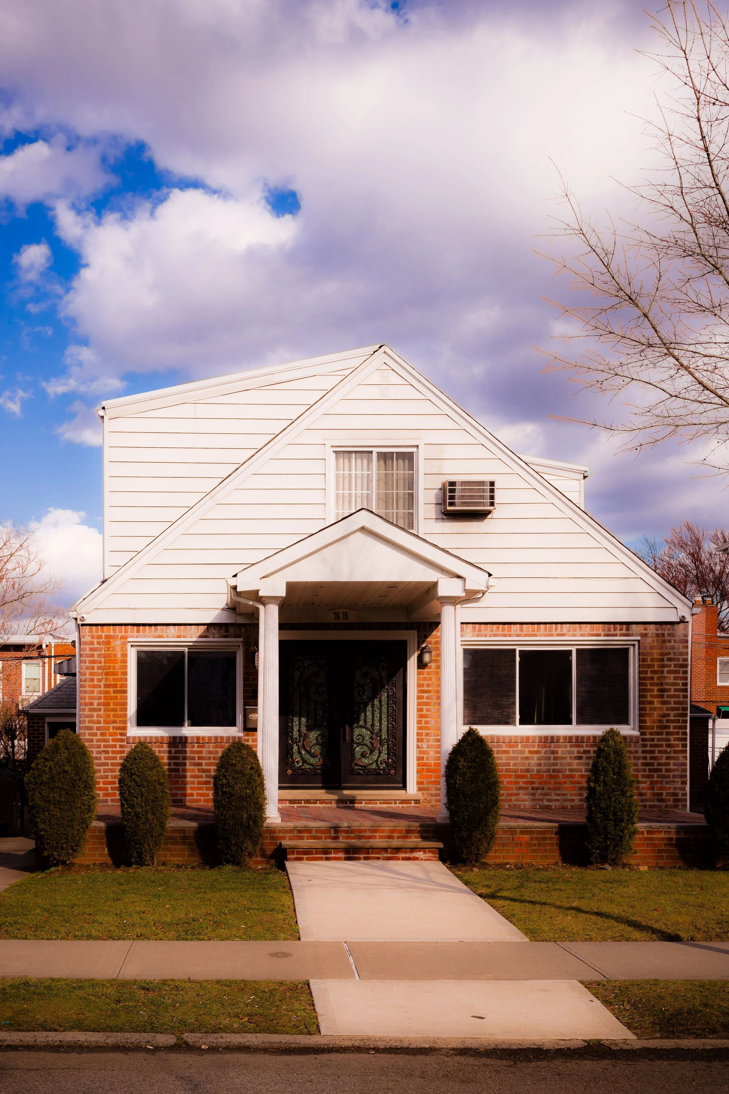 A two-story house with a brick lower level and white siding upper level, front porch with columns, and landscaped front yard with shrubs.
