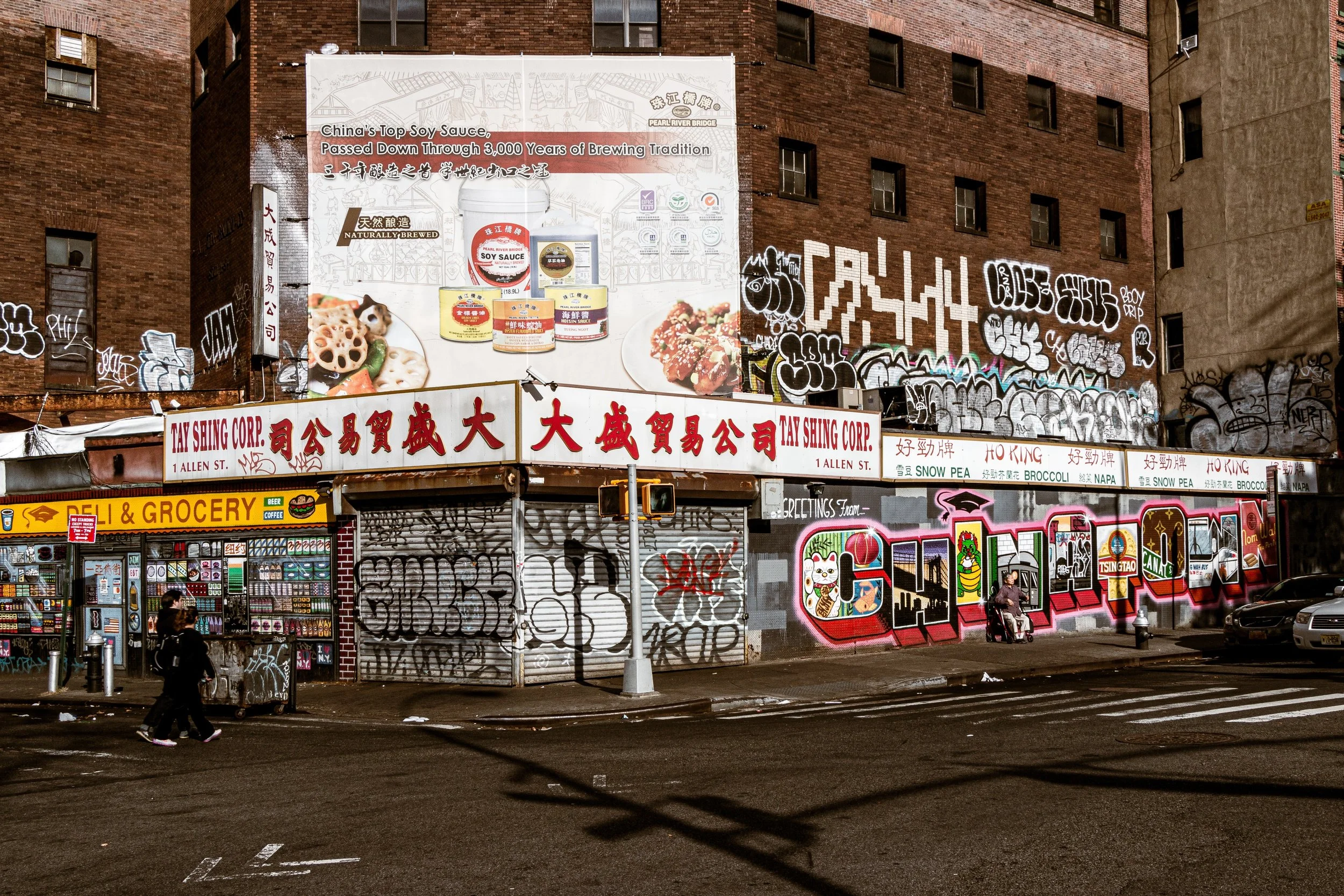 Street scene with graffiti-covered building, billboards, and a Chinatown sign on a storefront, with pedestrians walking and cars parked along the street at night.