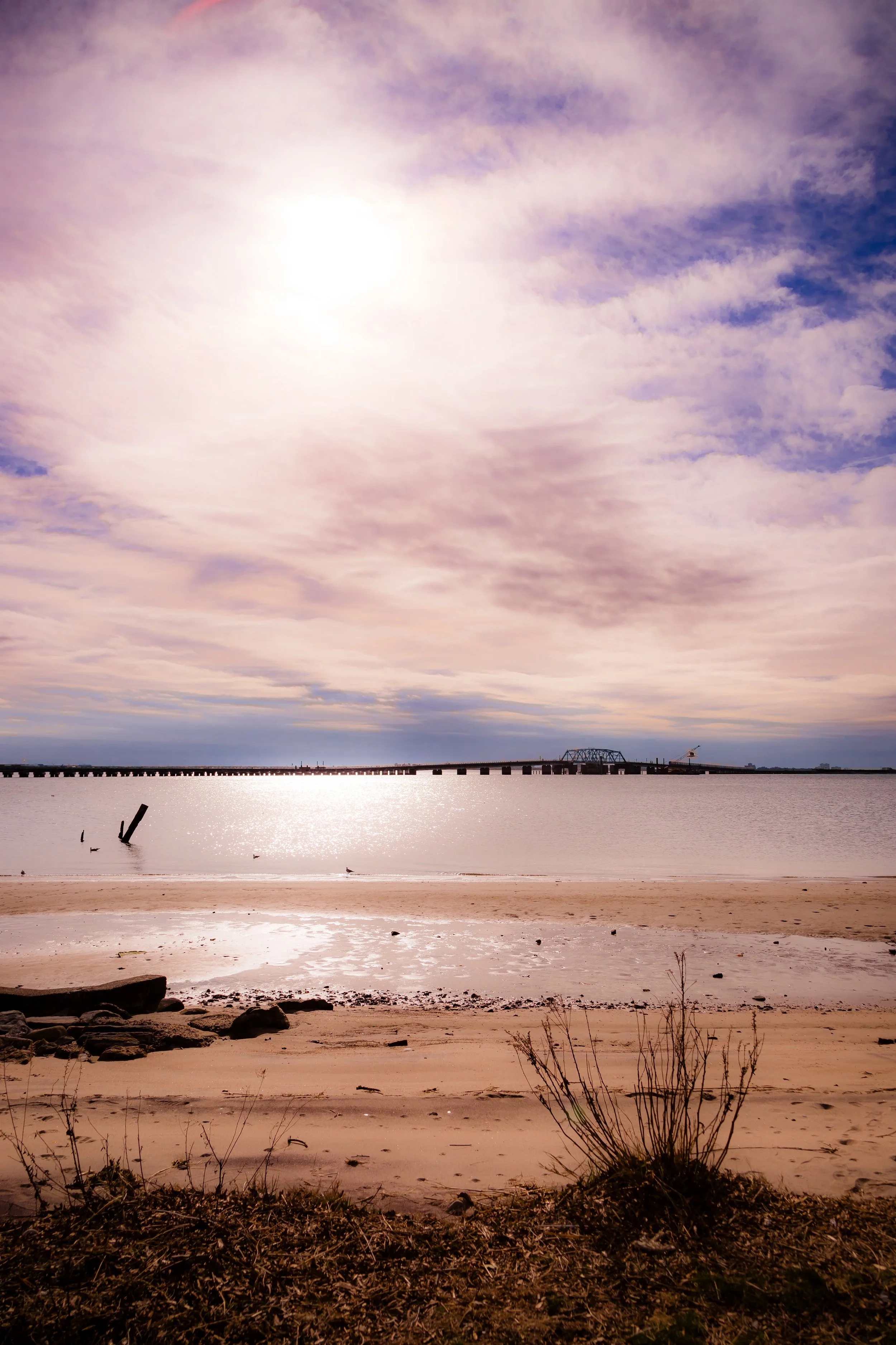A beach scene during daytime with a sandy shoreline, some rocks, a few sparse plants, and water reflecting the cloudy sky. In the background, there's a bridge across the water and a crane, with the sun partially obscured by clouds.