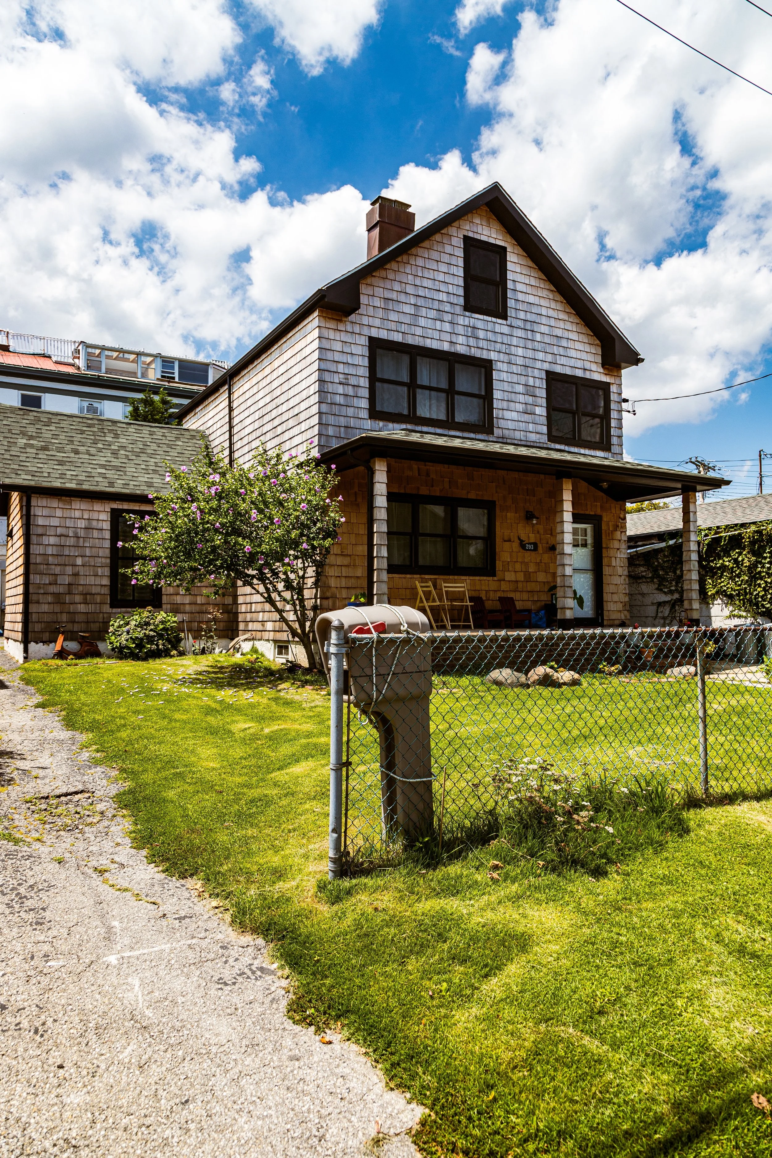 A two-story house with wooden shingles, black-framed windows, and a porch, surrounded by a green yard with a flowering tree, a chain-link fence, and a mailbox under a partly cloudy sky.