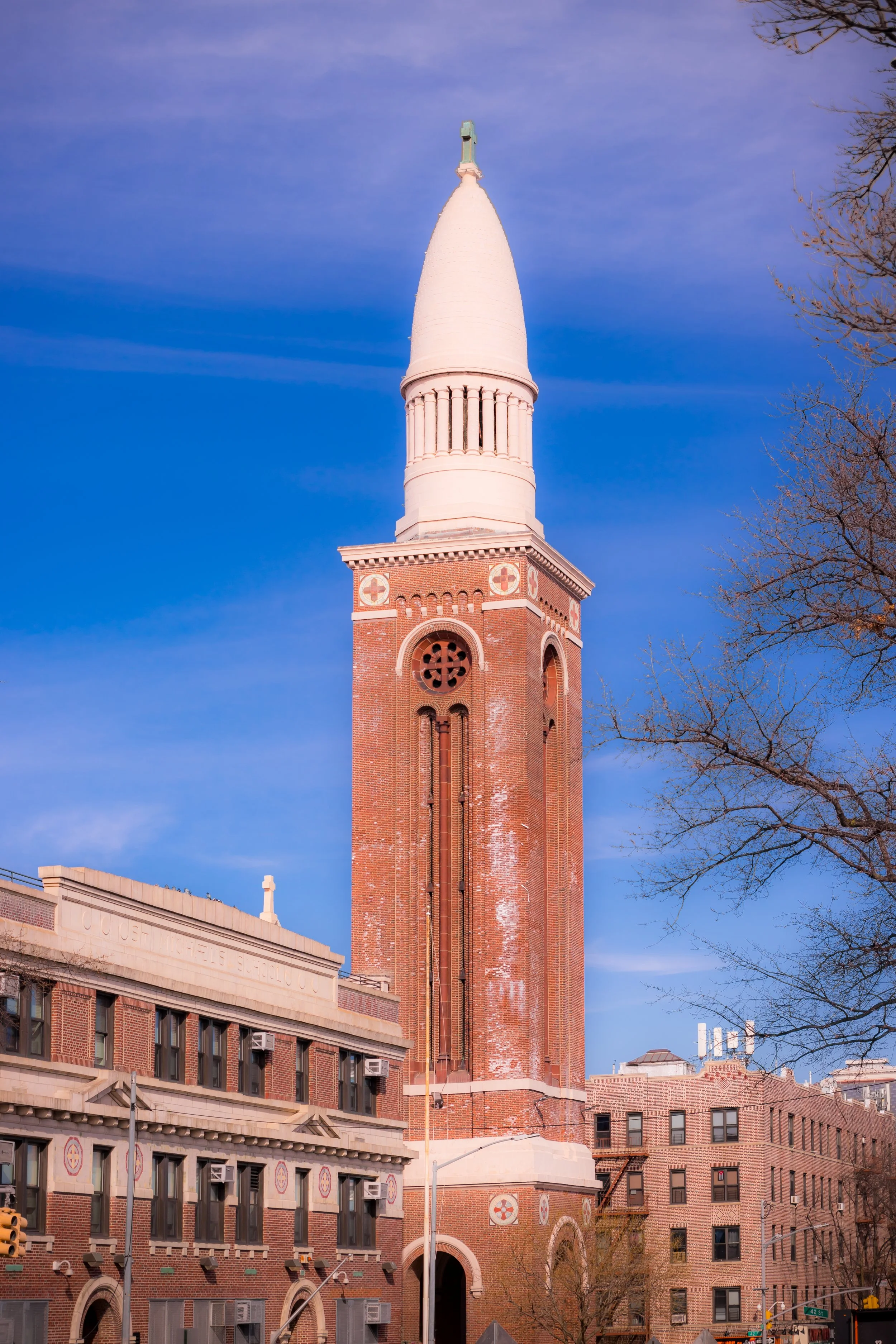 A tall brick clock tower with a white spire against a blue sky, situated next to multi-story brick buildings and leafless trees.