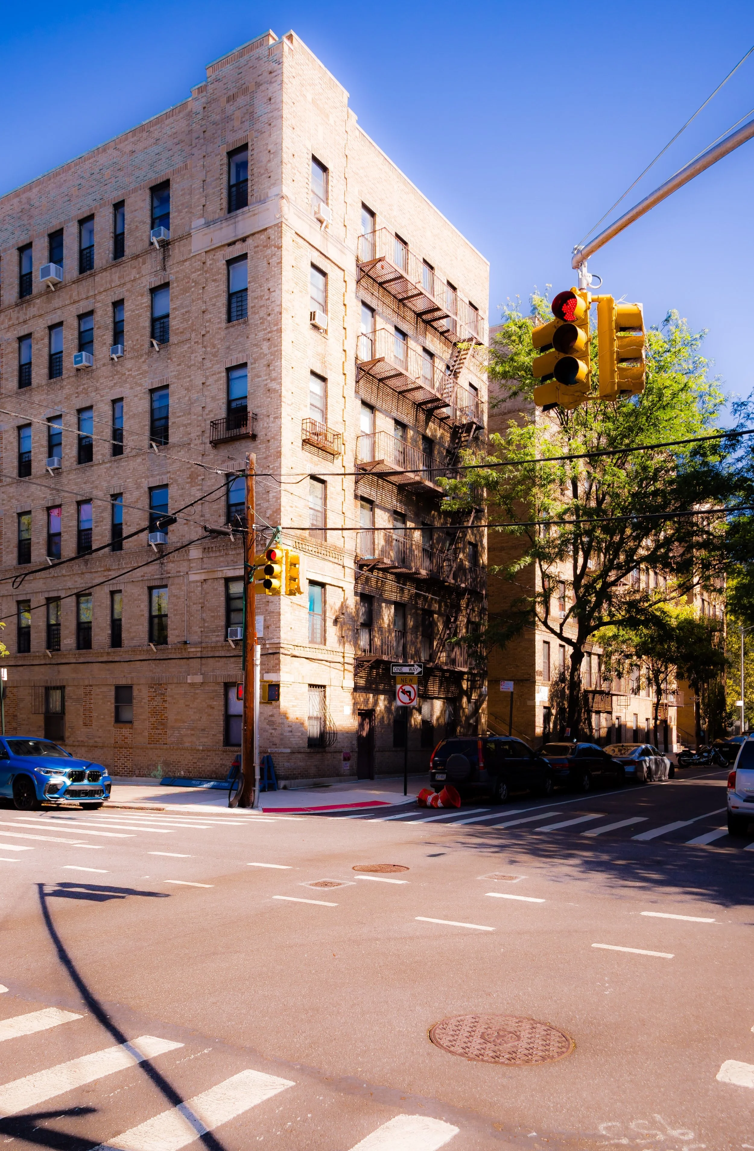 An urban street scene with a tall beige brick apartment building, traffic lights, cars parked on the side, and trees lining the street, under a bright blue sky.