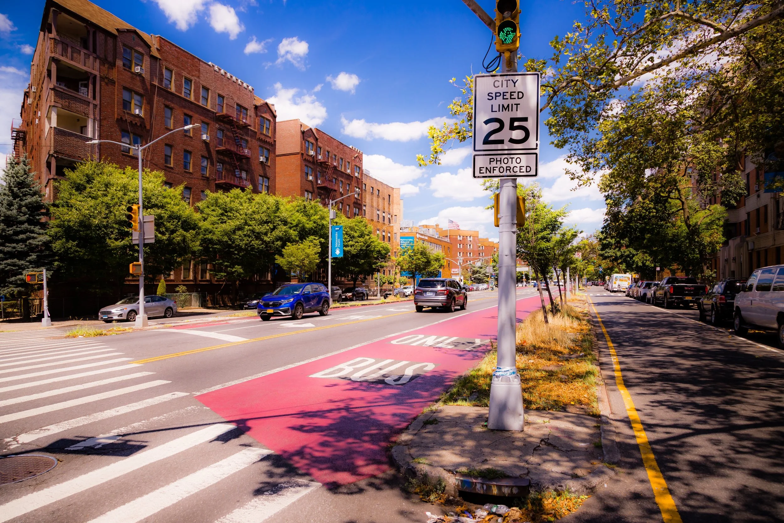 A city street with a clear sky, blue cars, and residential buildings. A green pedestrian signal and a speed limit sign indicating 25 mph with photo enforcement are visible. There are trees, a sidewalk, and a red bike lane marked with the words 'ONE BUS'.