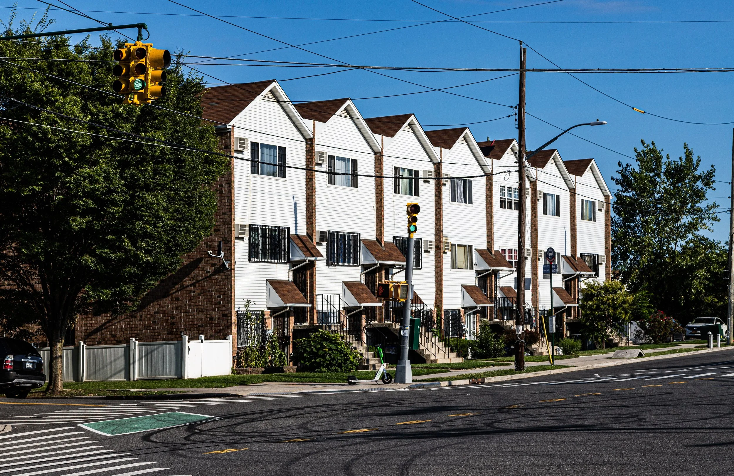 A row of white townhouses with brown roofs on a sunny day, with a traffic light and power lines in the foreground.