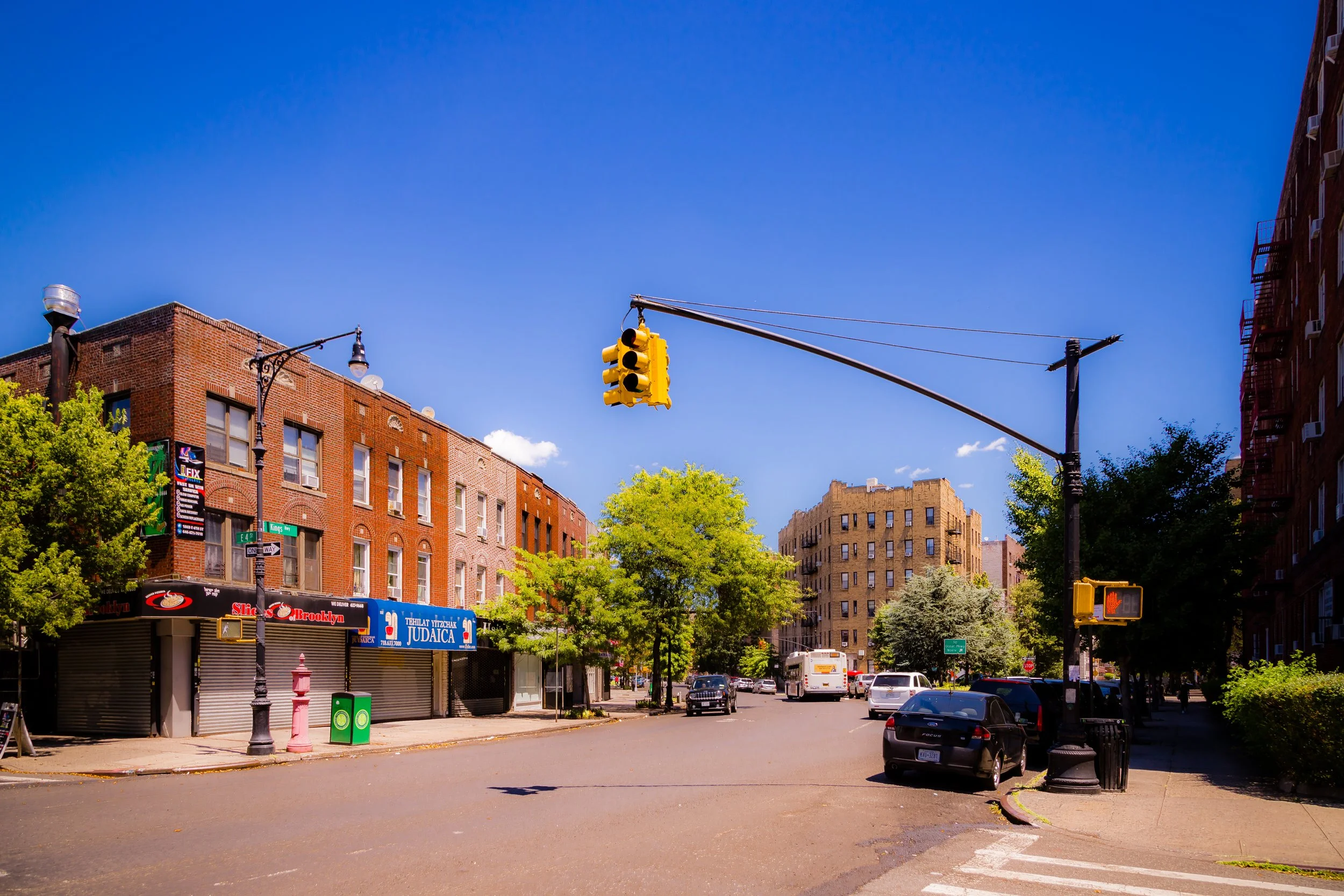City street scene with brick buildings, trees, parked cars, and a traffic light hanging over the intersection on a sunny day.