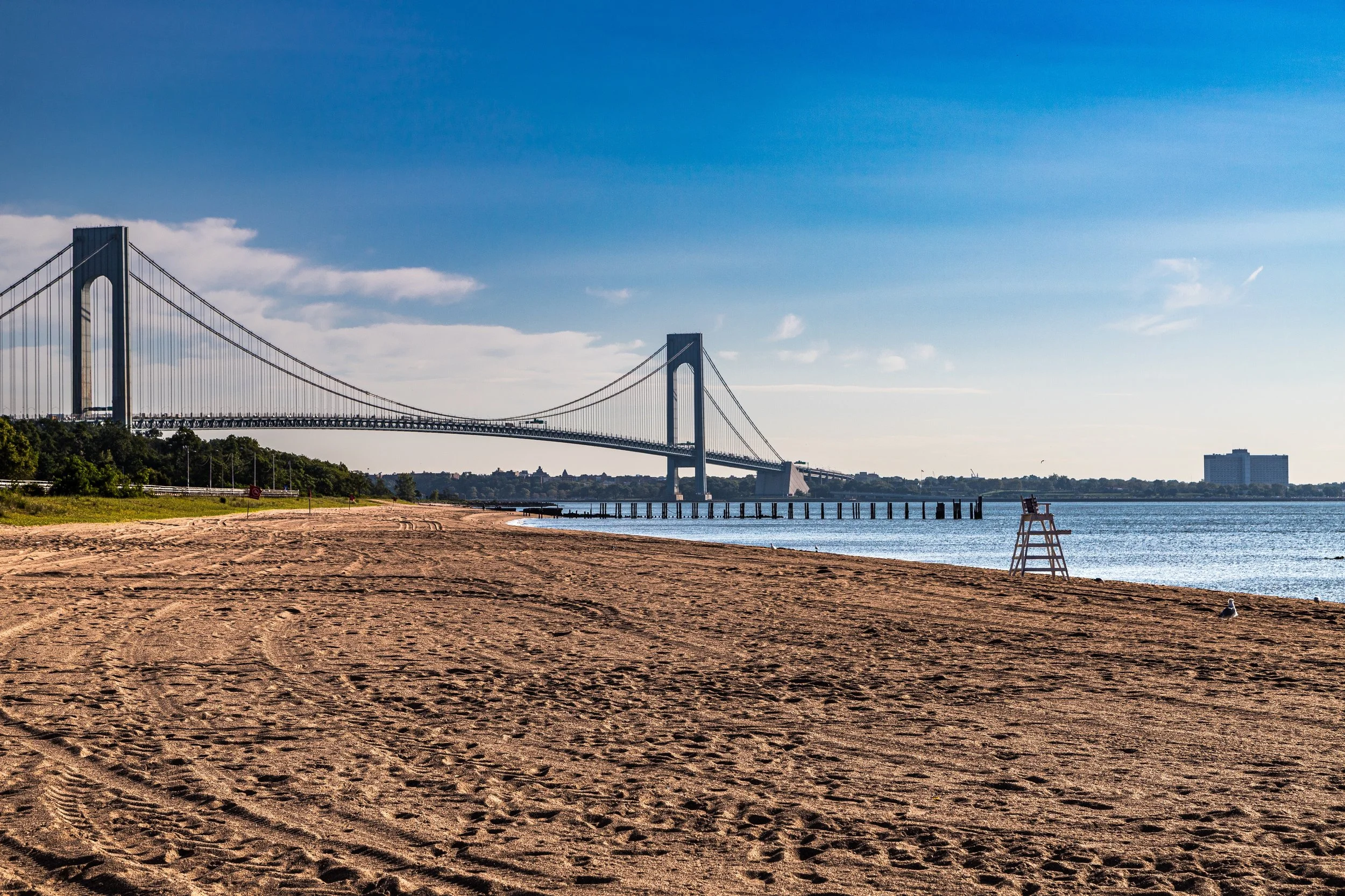 A sandy beach with footprints, a wooden lifeguard chair, and a metal pier, with the Verrazzano-Narrows Bridge in the background under a clear blue sky.