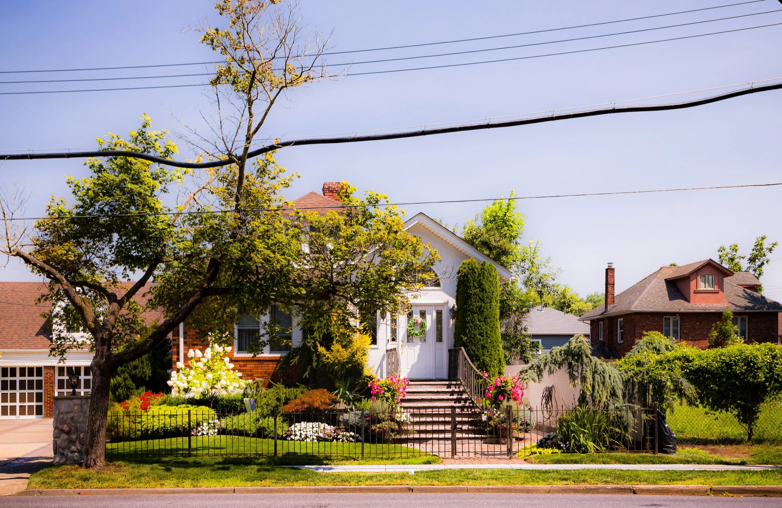 A residential house with a front yard, stairs leading to the door decorated with flowers and greenery, several trees, and neighboring houses visible in the background, under a clear sky.