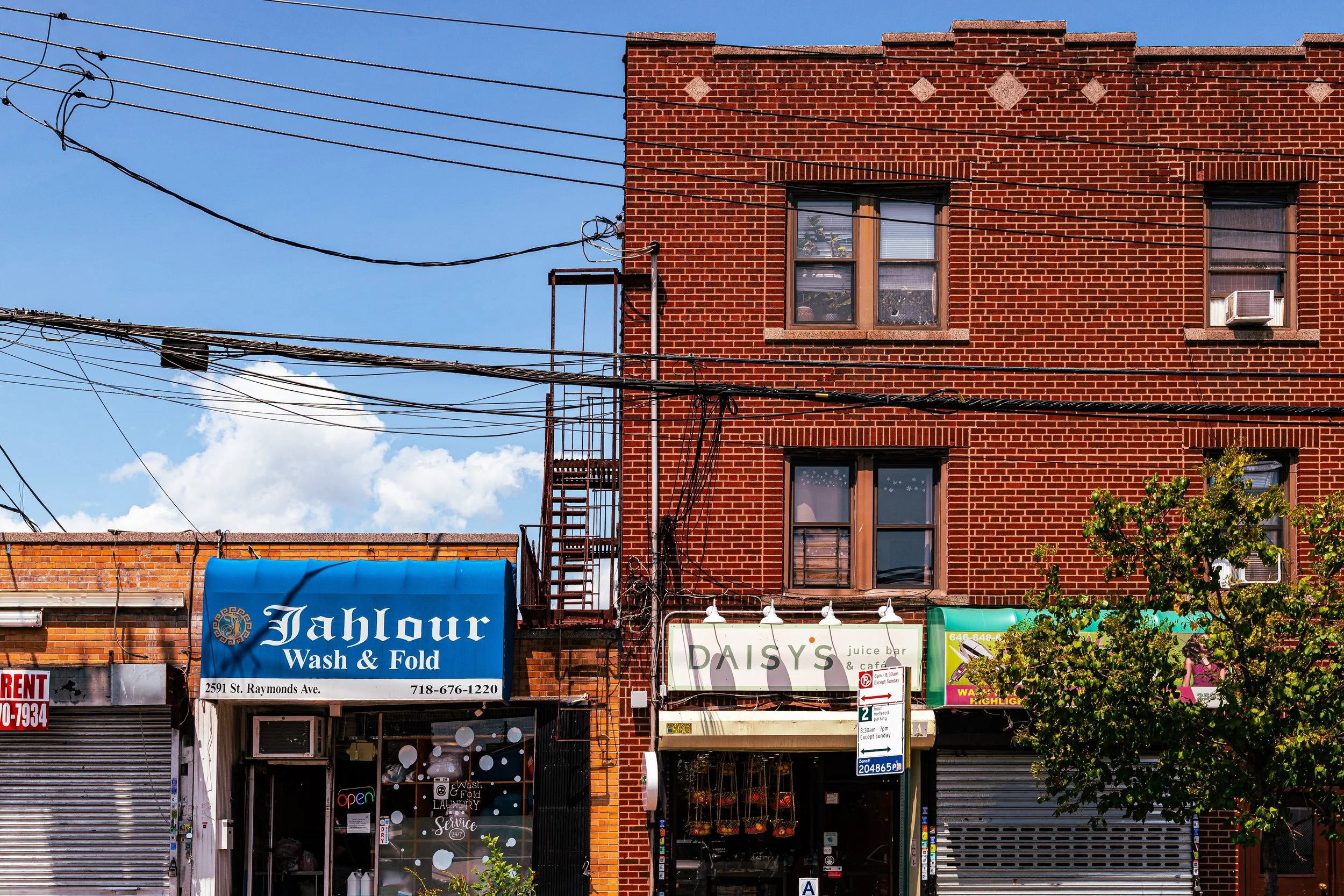 Three storefronts on a city street with three-story brick building above. The middle storefront is 'Daisy's Juice Bar & Cafe' with hanging flower baskets. The left storefront is 'Jahlour Wash & Fold' with a blue sign, and the right storefront has a closed metal shutter. Power lines cross the blue sky with some clouds. A small tree is in front of the right storefront.