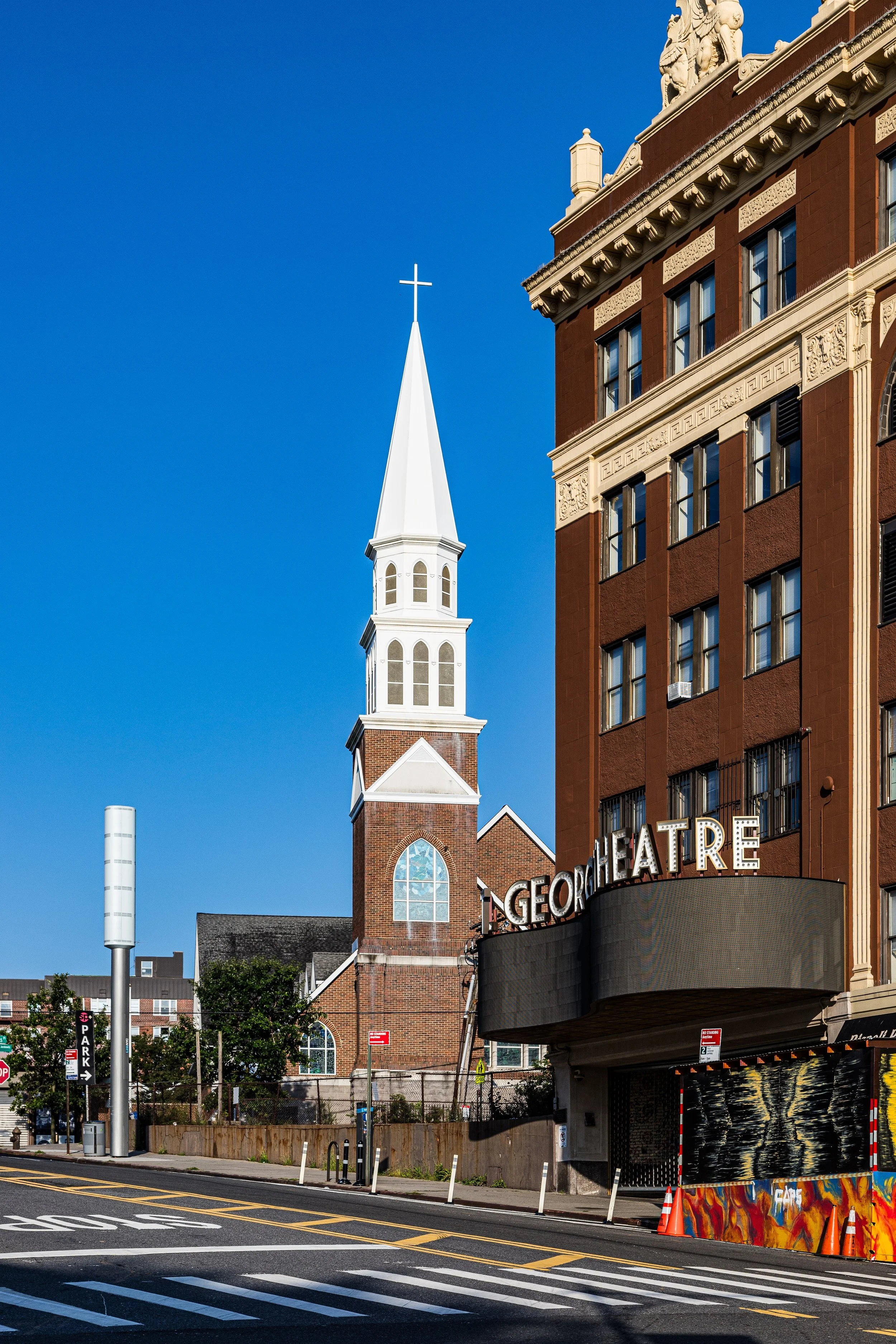 Street view featuring a church with a tall steeple and cross, adjacent to a historic theater building on a sunny day.
