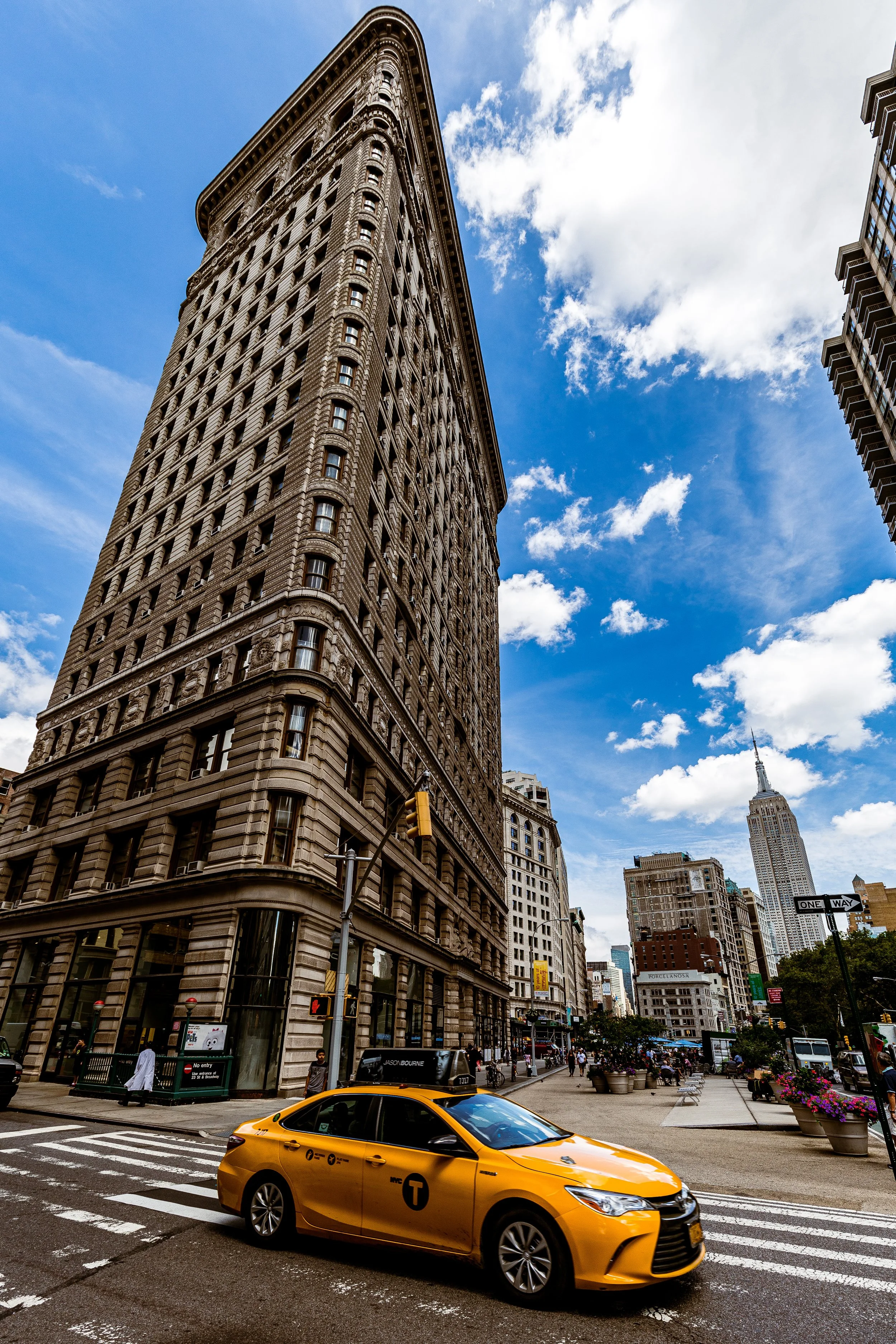 Street scene in New York City with the Flatiron Building, a yellow NYC taxi, pedestrians, and the Empire State Building in the background on a partly cloudy day.