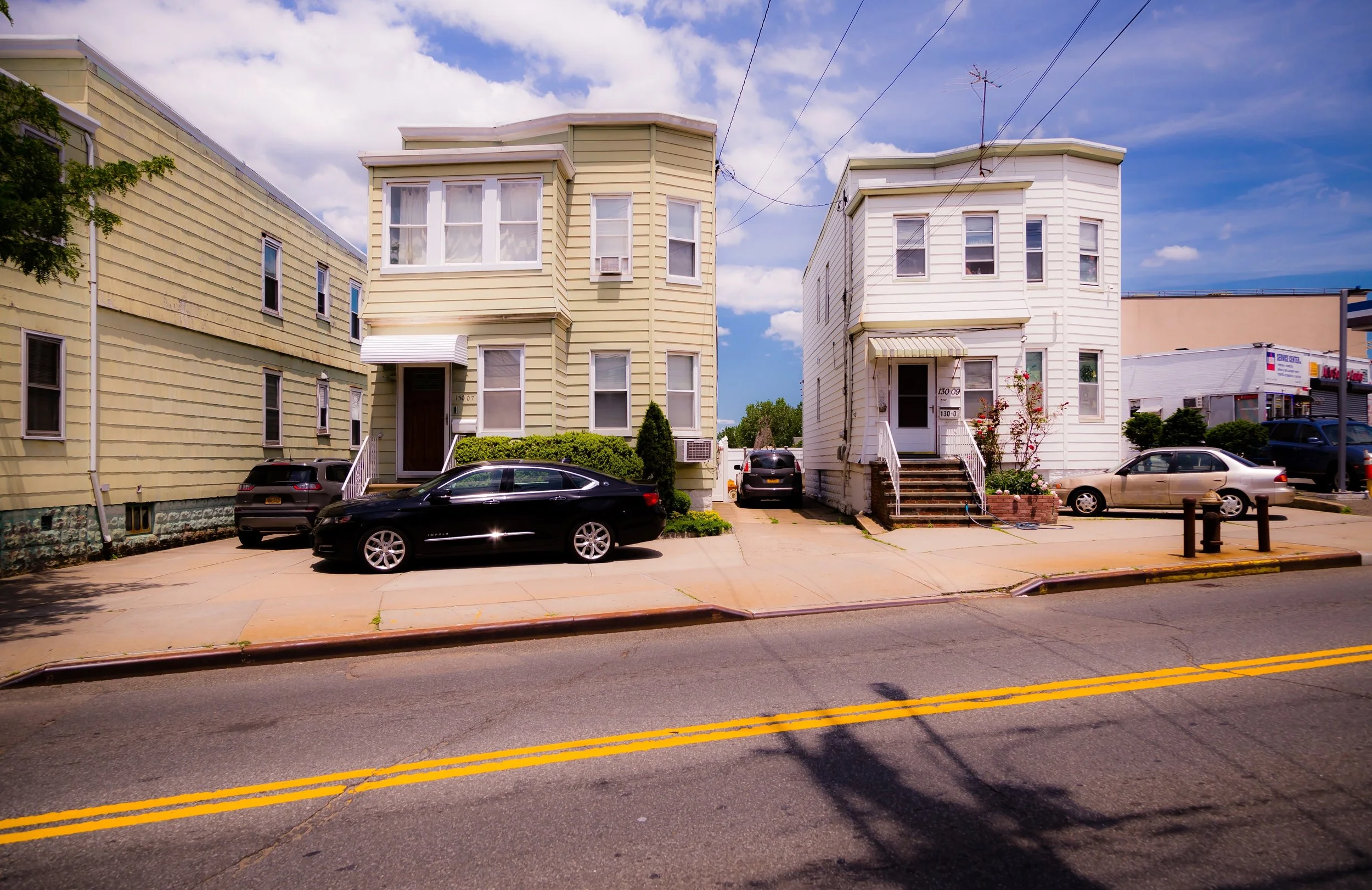 Two pastel-colored houses with small front yards, parked cars on the street, and a clear sky with a few clouds.