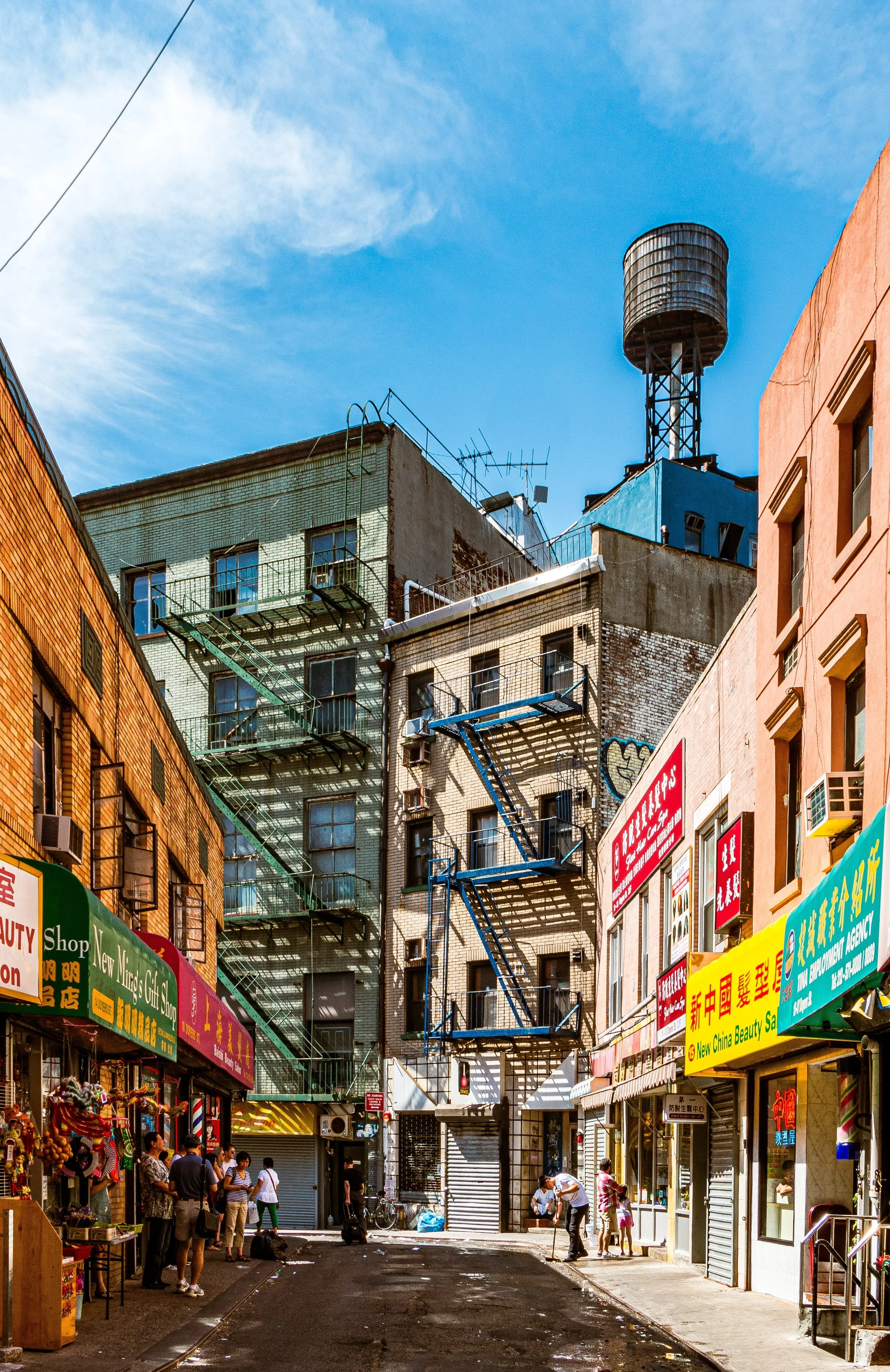 City street scene with buildings, fire escapes, colorful signs in Chinese, and people walking and cleaning on the sidewalk.
