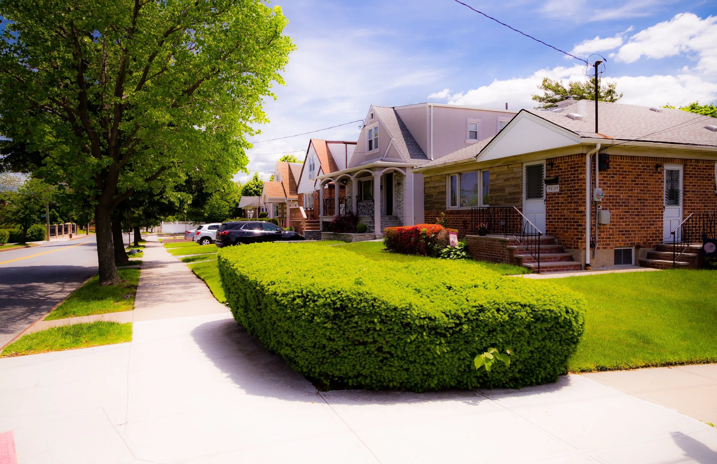 A row of houses with front yards and parked cars along a tree-lined street on a sunny day with a blue sky and white clouds.