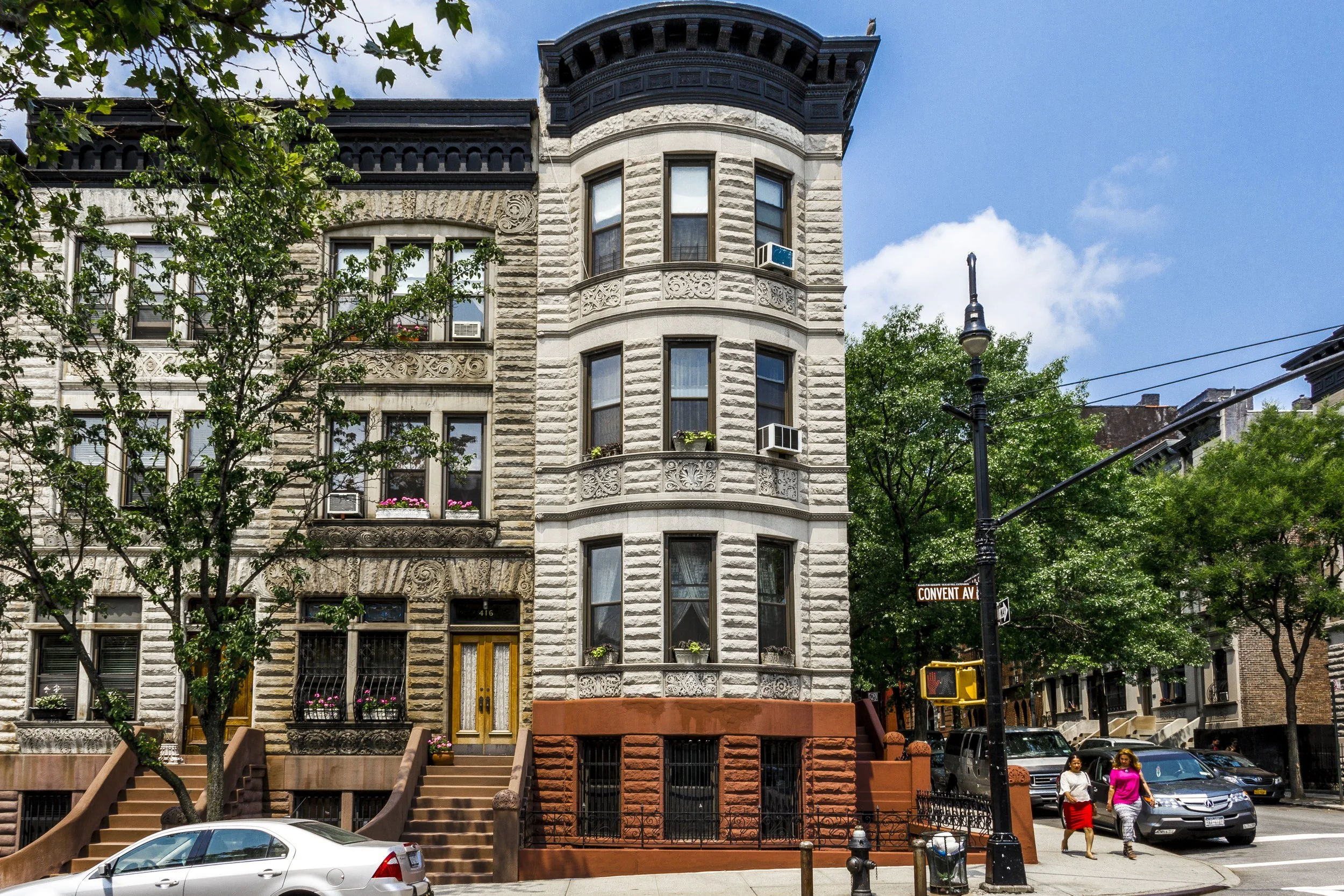 A row of historic brownstone buildings on a city street, with stairs leading up to their entrances, trees lining the sidewalk, cars parked alongside, and two women walking on the sidewalk on a sunny day.