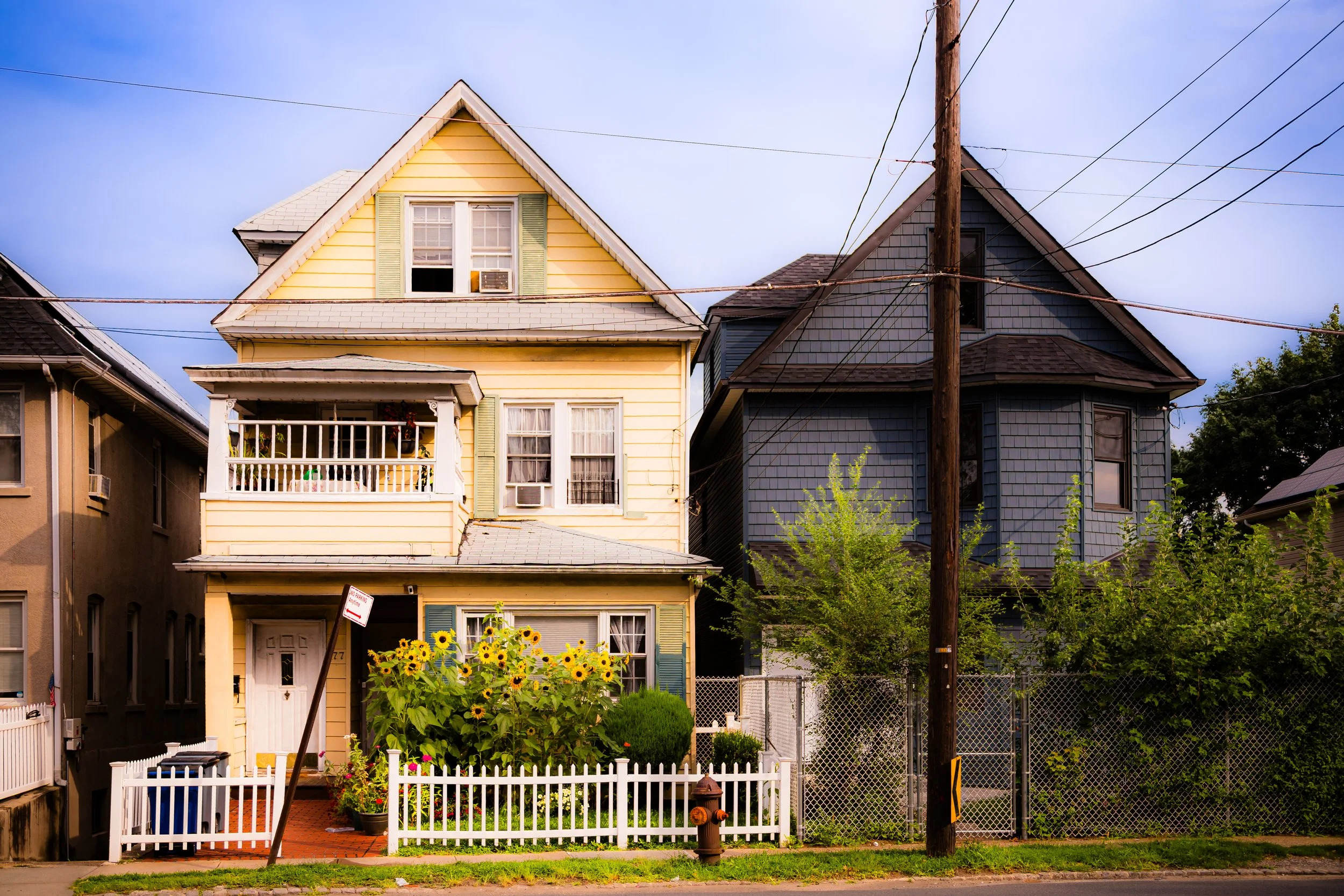 A yellow three-story house with a porch and sunflower garden on the front, next to a dark blue house with a fenced yard, power pole, and greenery.