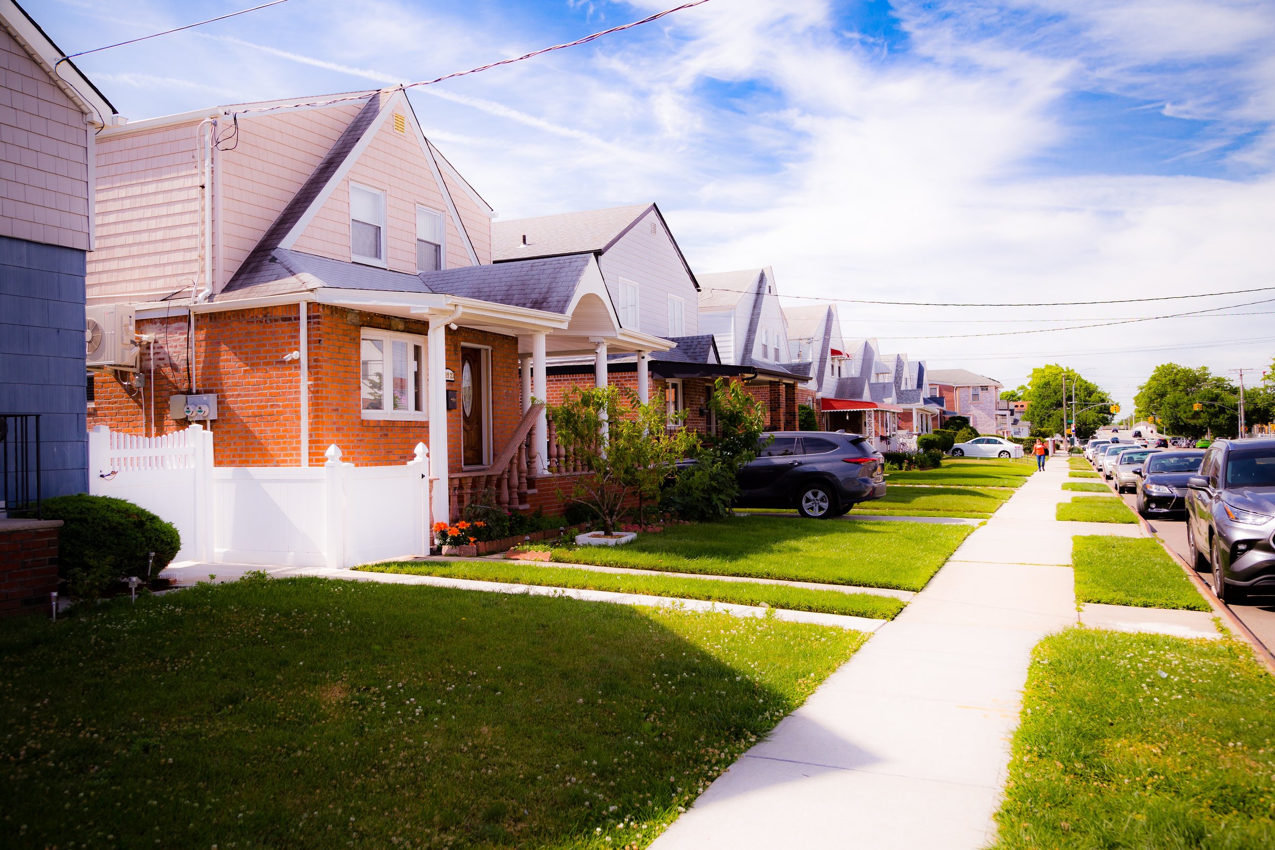 A row of suburban houses with front yards, parked cars, and a sidewalk, under a partly cloudy sky.