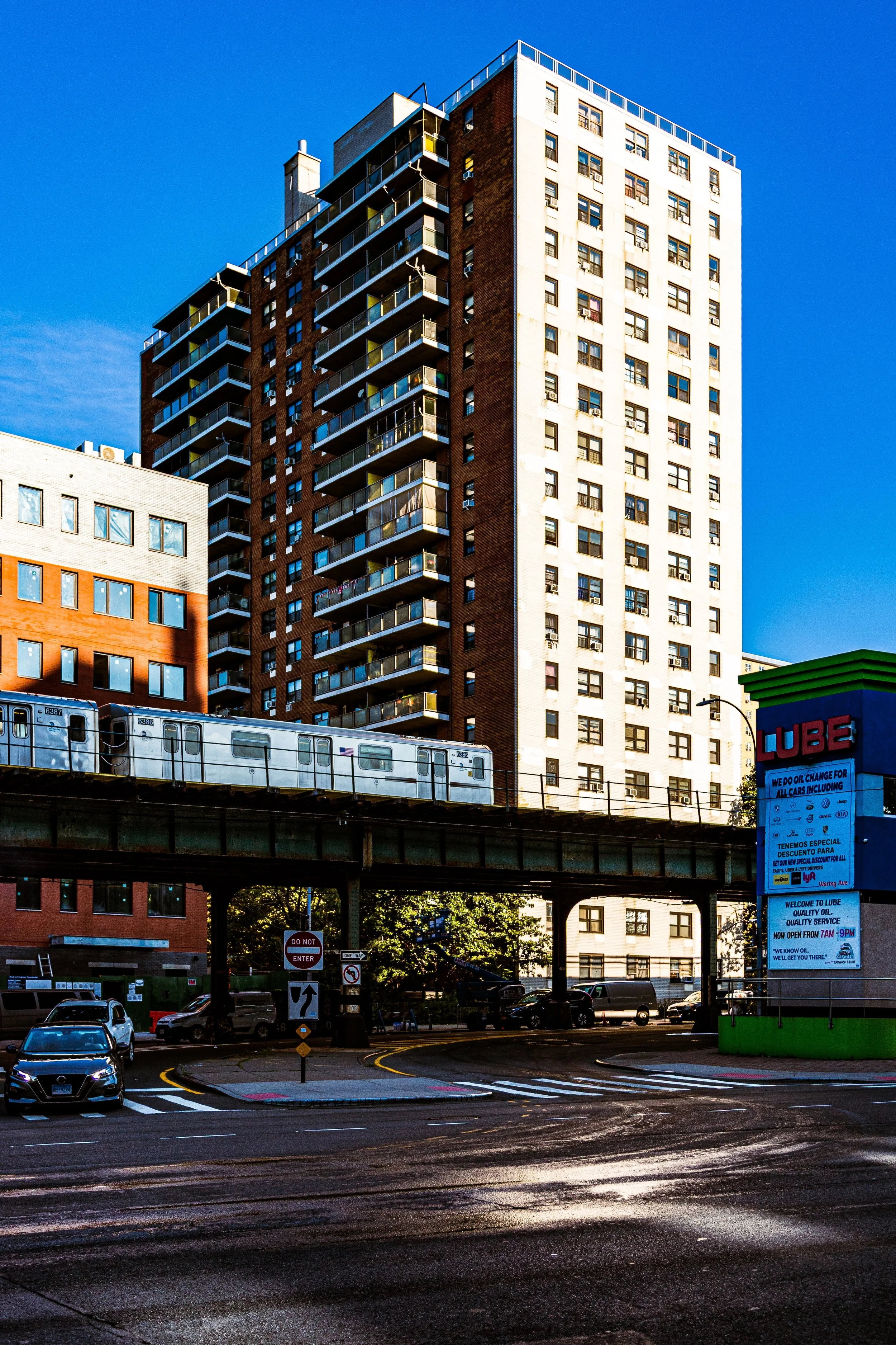 Tall apartment building with balconies under a blue sky, with a train passing on an elevated track, and cars parked below on the street.