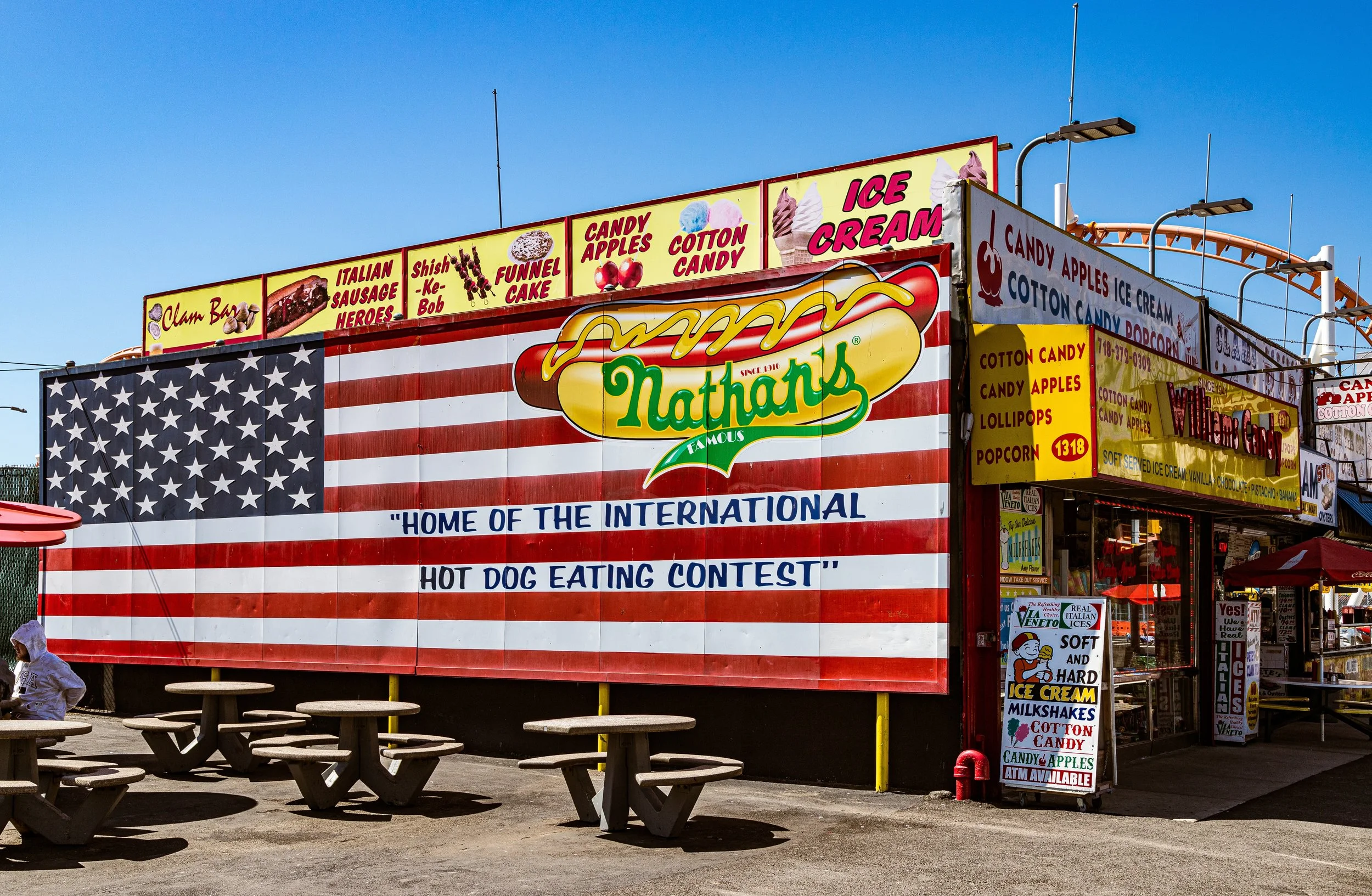 Exterior of Nathan's famous hot dog stand with a large American flag mural, colorful signs advertising ice cream, candy apples, cotton candy, and hot dogs, and a few picnic tables outside.
