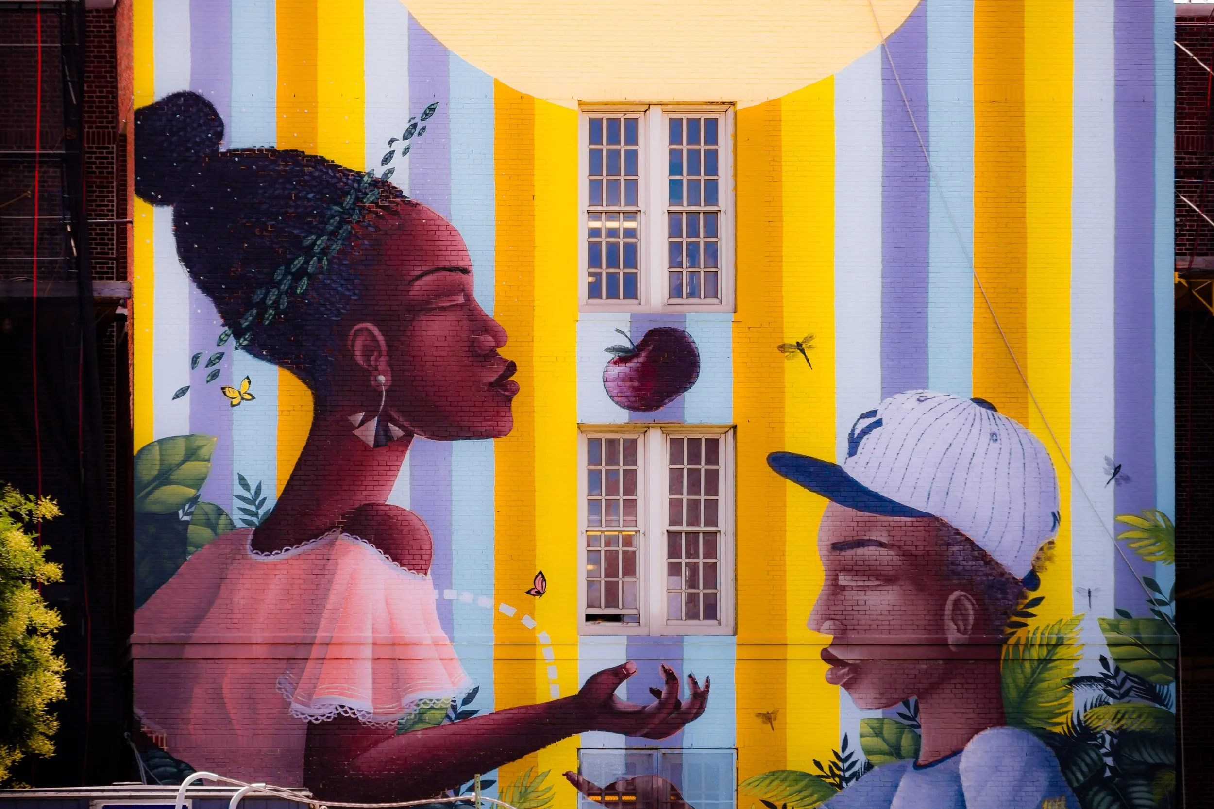 Colorful mural featuring two women facing each other, one with a large bun hairstyle and the other wearing a baseball cap, surrounded by natural elements like leaves, butterflies, and a fruit in the background.