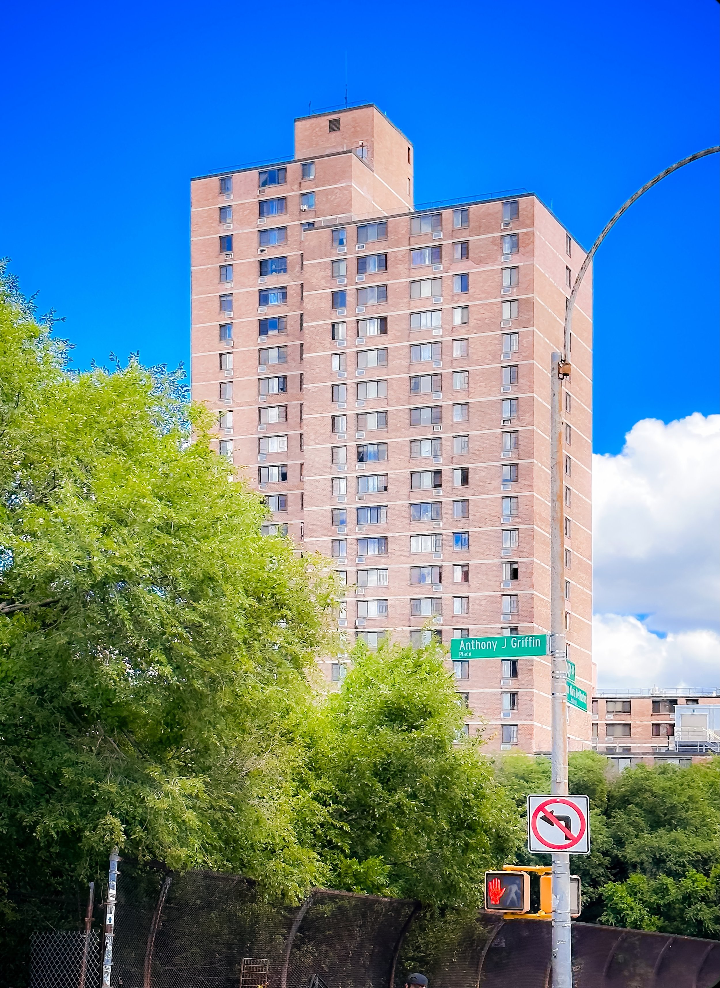 A tall pink brick residential building with many windows, a street sign reading Anthony J Griffin Place, a no left turn traffic sign, and a pedestrian signal showing a red hand, with green trees and a bright blue sky in the background.