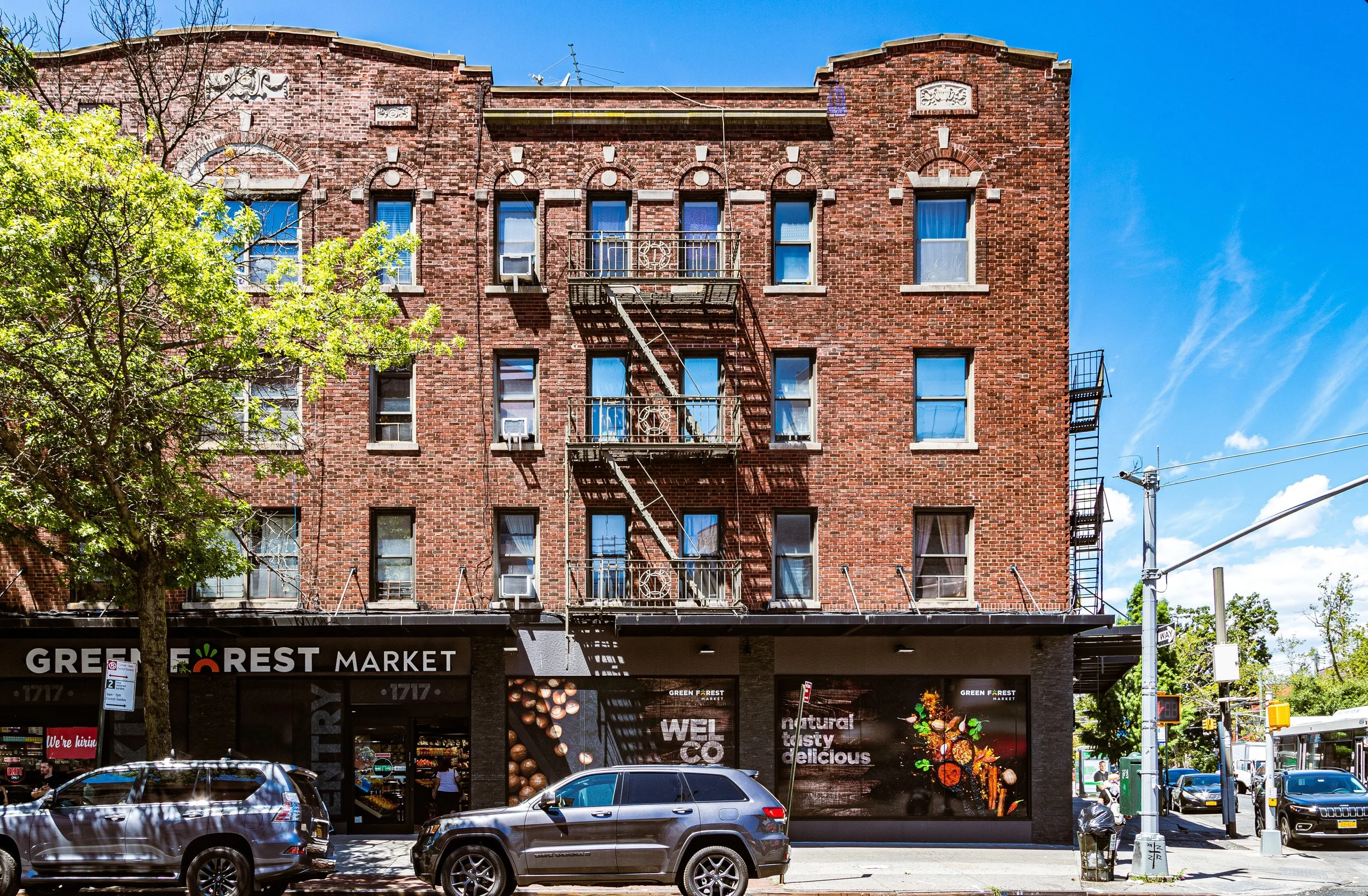 A brick building with a tree in front, a fire escape on the facade, and a storefront with signs for Green Forest Market. Cars are parked on the street, and a bus is visible at the corner, with a blue sky overhead.