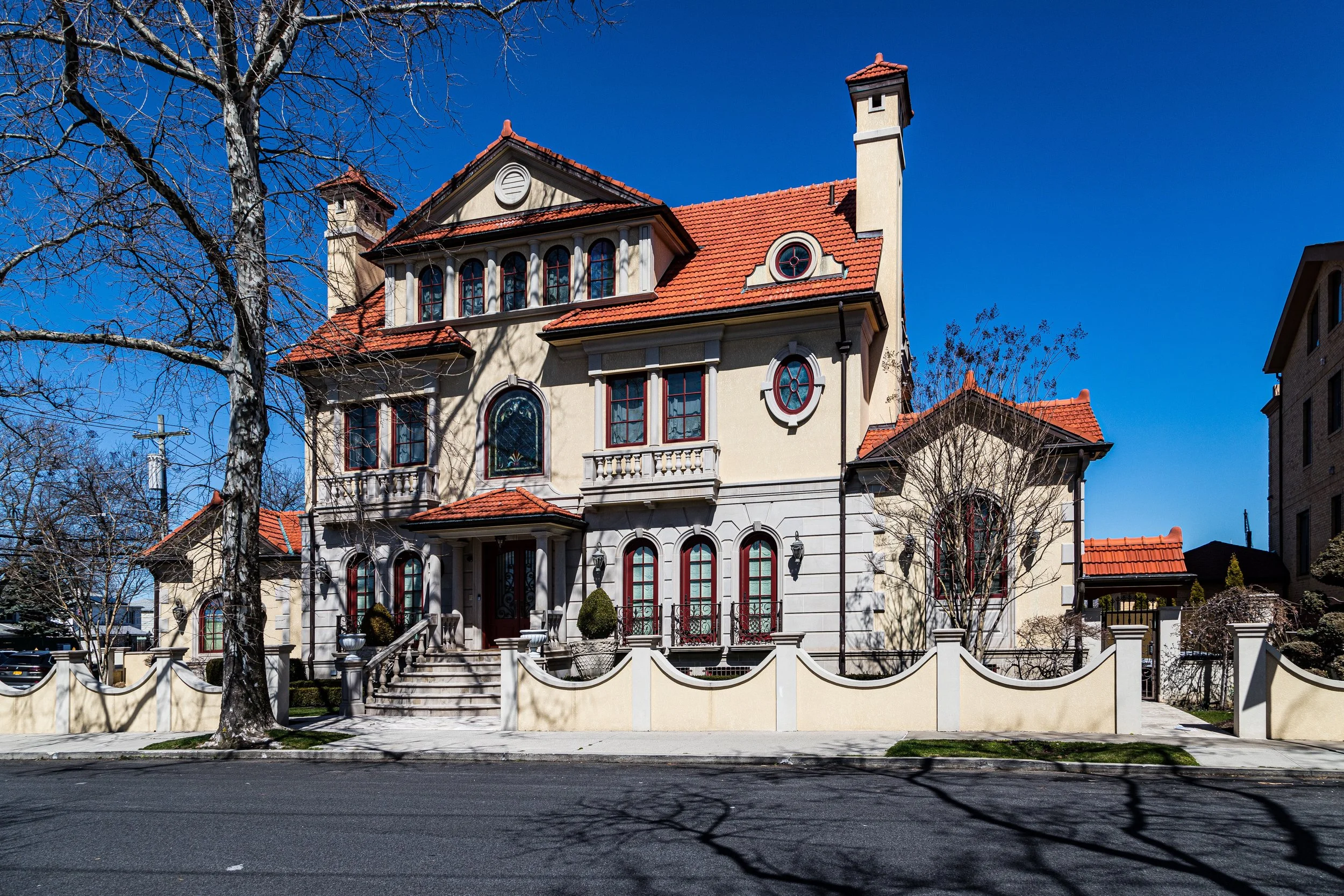 A large house with a beige exterior, red tile roof, and multiple windows, surrounded by a decorative cream-colored fence. Tall leafless trees are in front, with a bright blue sky in the background.